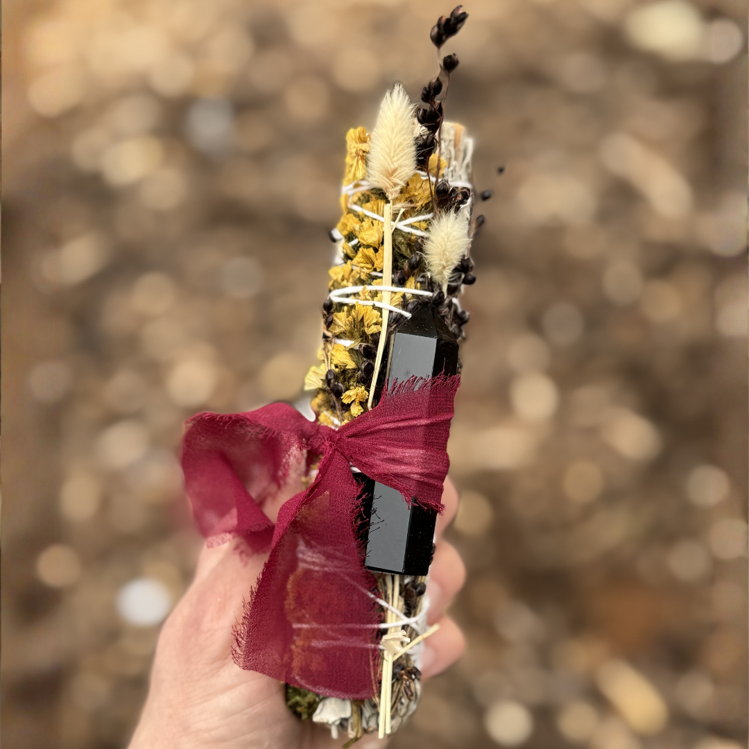 Handcrafted 6-inch White Sage smudge stick wrapped with a Black Obsidian crystal point, yellow sinuata flowers, bunny tails, broom corn and a burgundy ribbon. Held in hand outdoors with a soft natural background.
