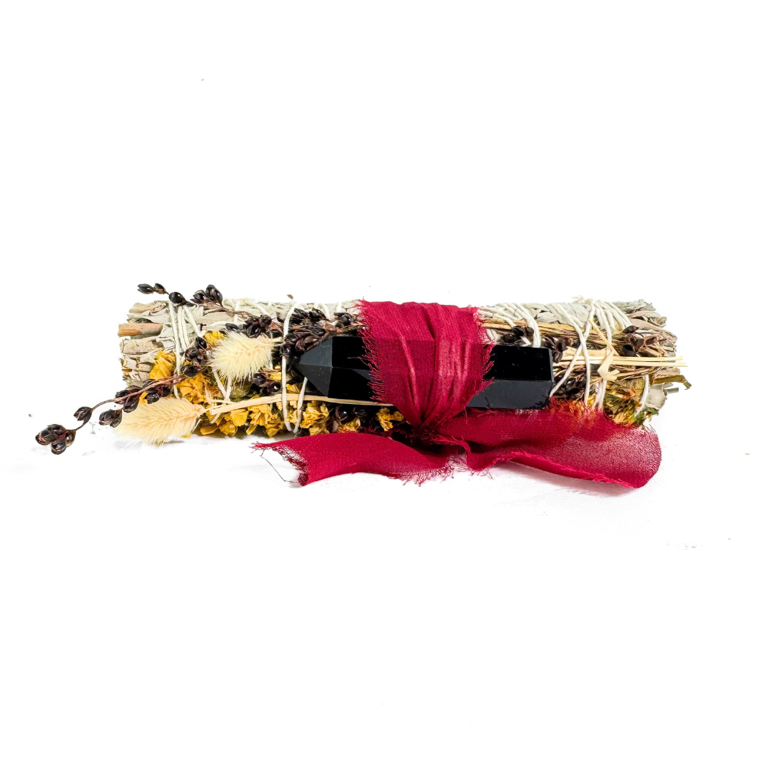 Handcrafted 6-inch White Sage smudge stick wrapped with a Black Obsidian crystal point, yellow sinuata flowers, bunny tails, broom corn and a burgundy ribbon. Held in hand outdoors with a white natural background.