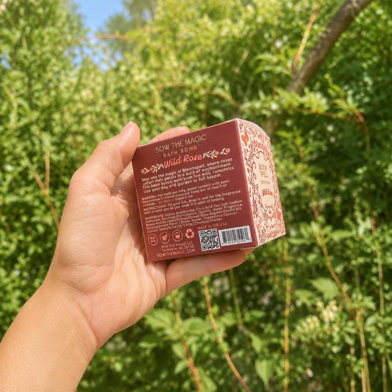 Hand holding the back of the Sow the Magic Bloom Spell Wild Rose bath bomb box in a lush summer garden with softened natural sunlight, highlighting the product details against vibrant green foliage and a dreamy botanical backdrop.