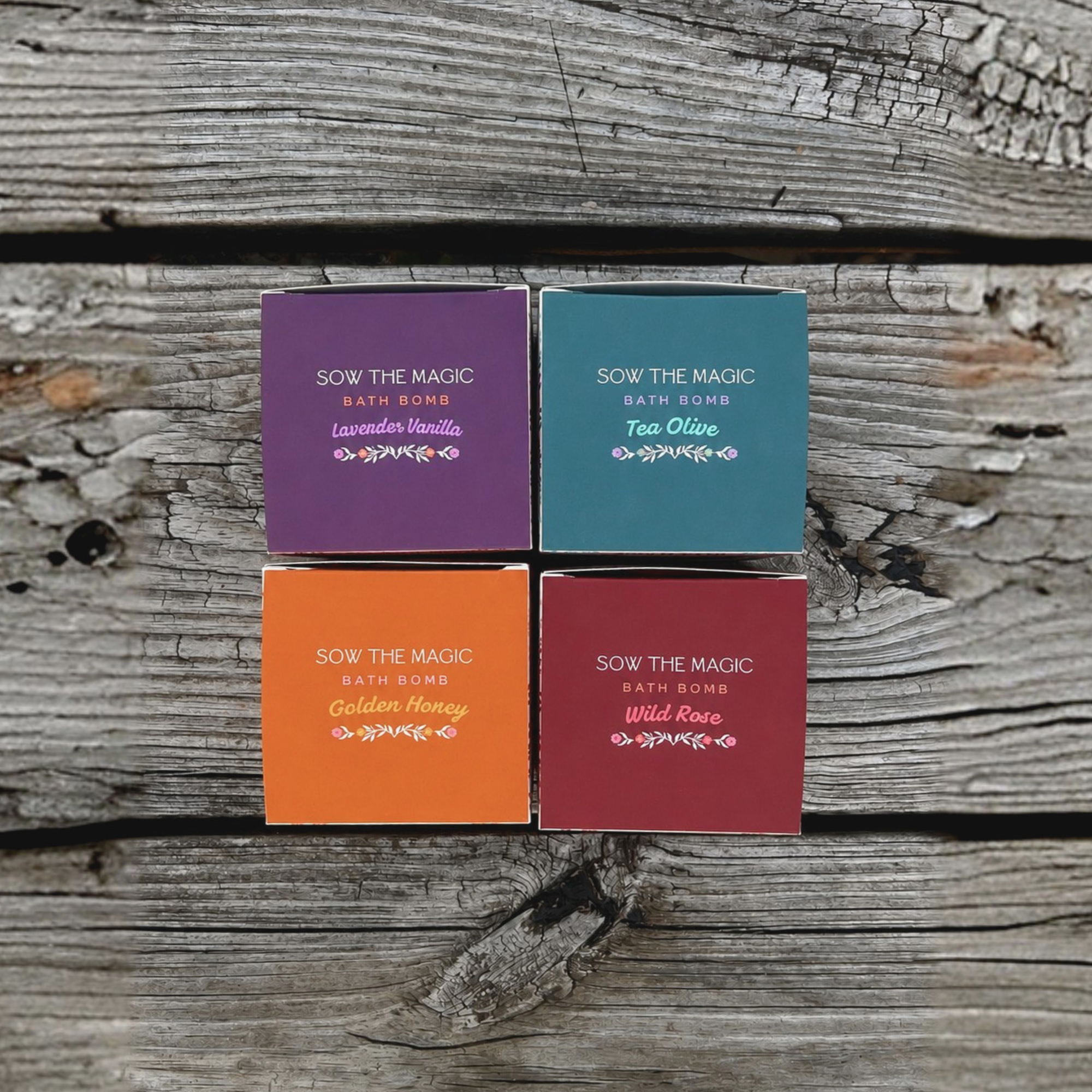 Top view of the Bloomspell botanical bath bomb collection featuring Tea Olive, Wild Rose, Lavender Vanilla and Golden Honey gift boxes arranged on a rustic wooden surface.