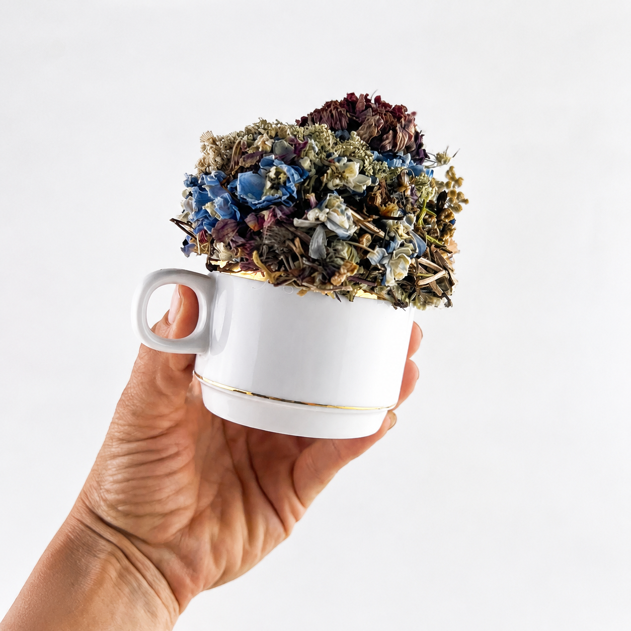 Handmade white teacup arrangement with blue, burgundy and cream dried farm botanicals held in a hand.