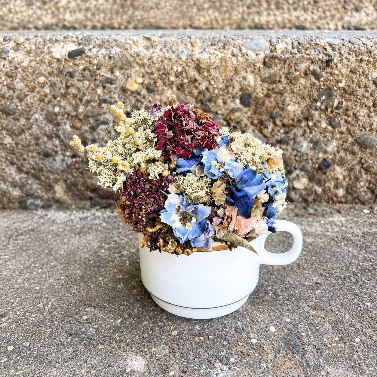 Blue, cream and burgundy handmade teacup flower arrangement displayed on an outdoor stone step.