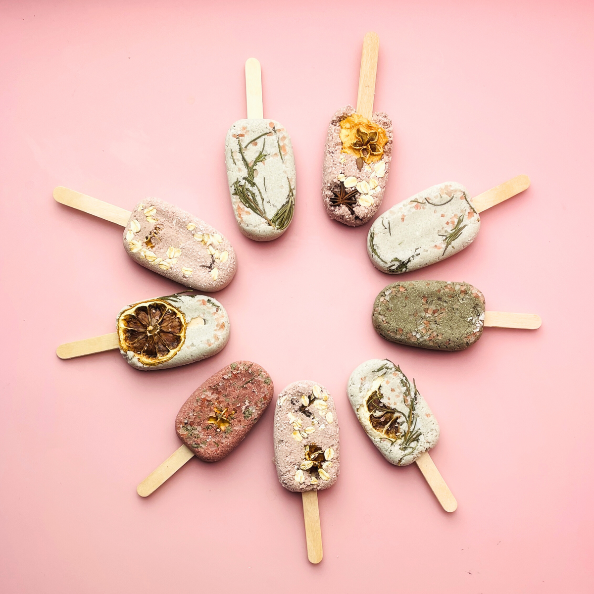 Assorted popsicle-shaped bath bombs with dried flowers and citrus on pink background
