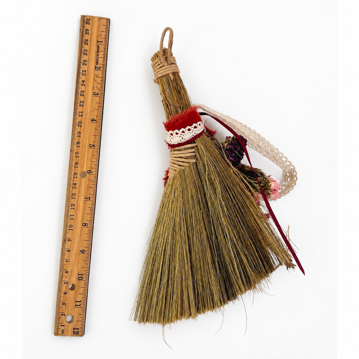 Back view of a handmade straw botanical broom beside a 12-inch ruler, showing its natural bristles, wrapped handle and ribbon details.