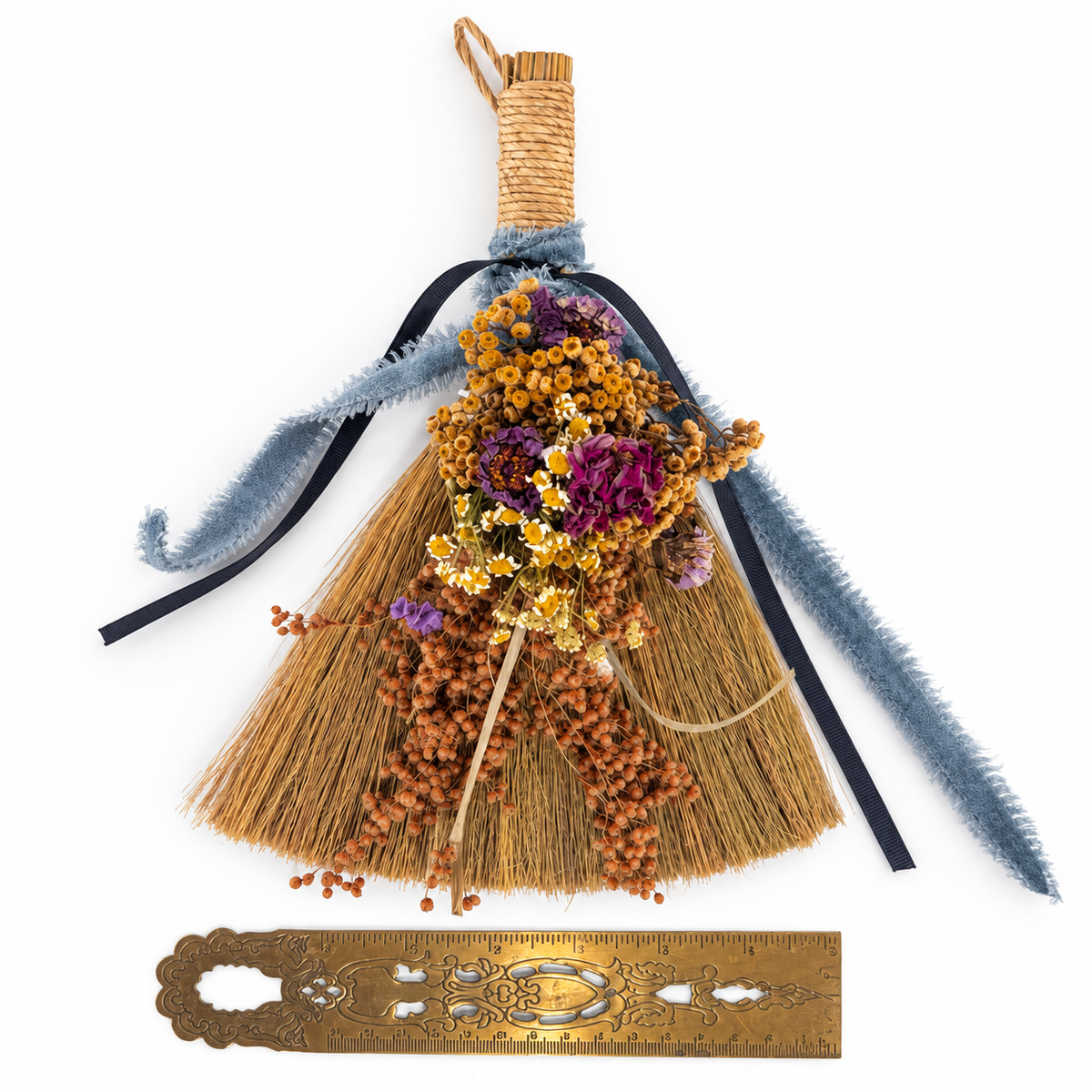 Handmade botanical broom with dried purple and yellow flowers, natural straw, and blue ribbons displayed beside an antique brass ruler for size reference.
