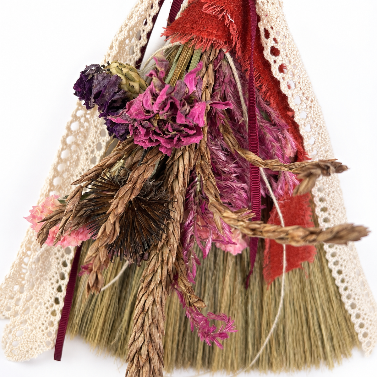 Close-up of dried pink flowers, natural wheat, textured botanicals, red ribbon and vintage-style cream lace on a handmade broom.