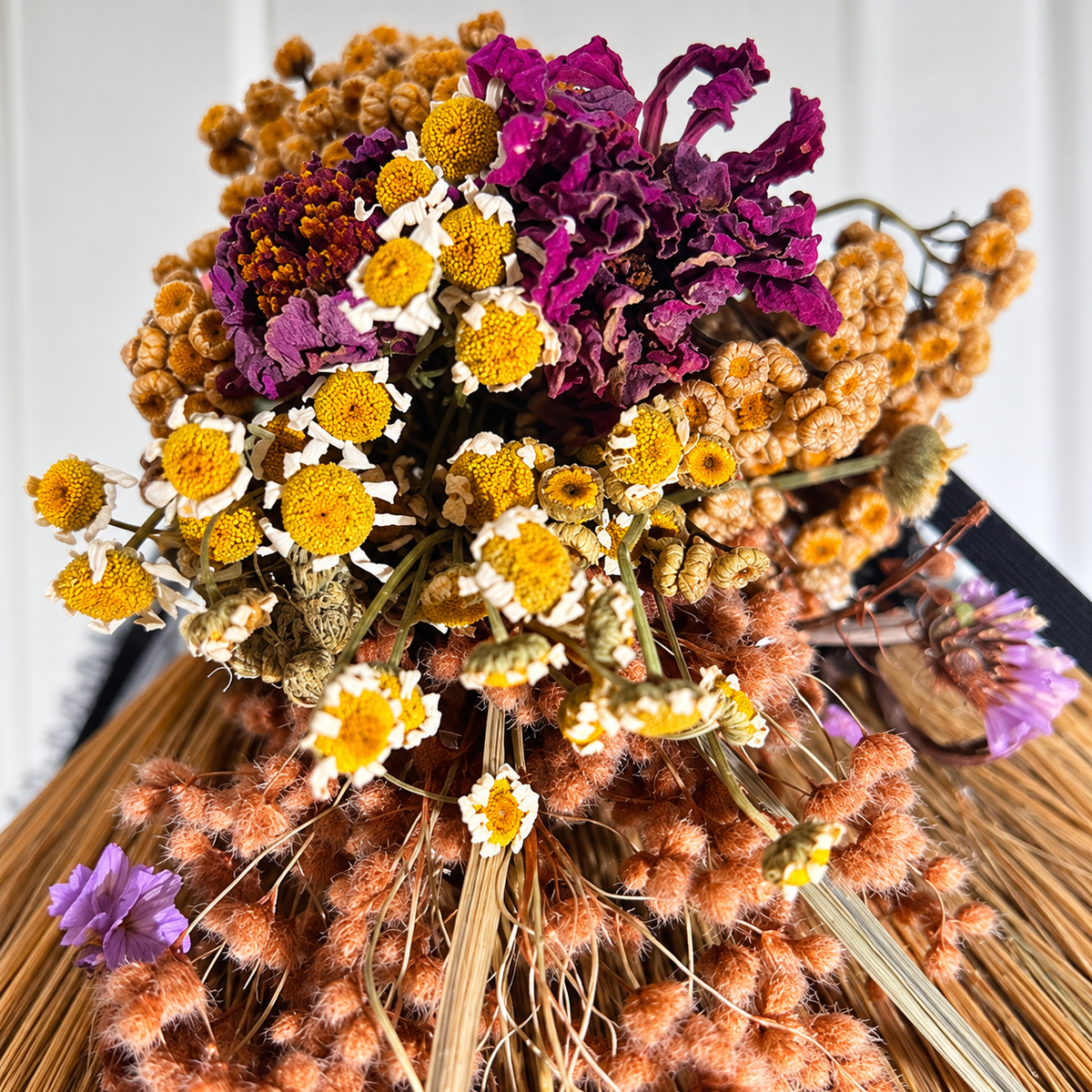 Close-up of dried purple flowers, yellow chamomile blooms, and textured botanicals arranged on a handcrafted decorative straw broom.