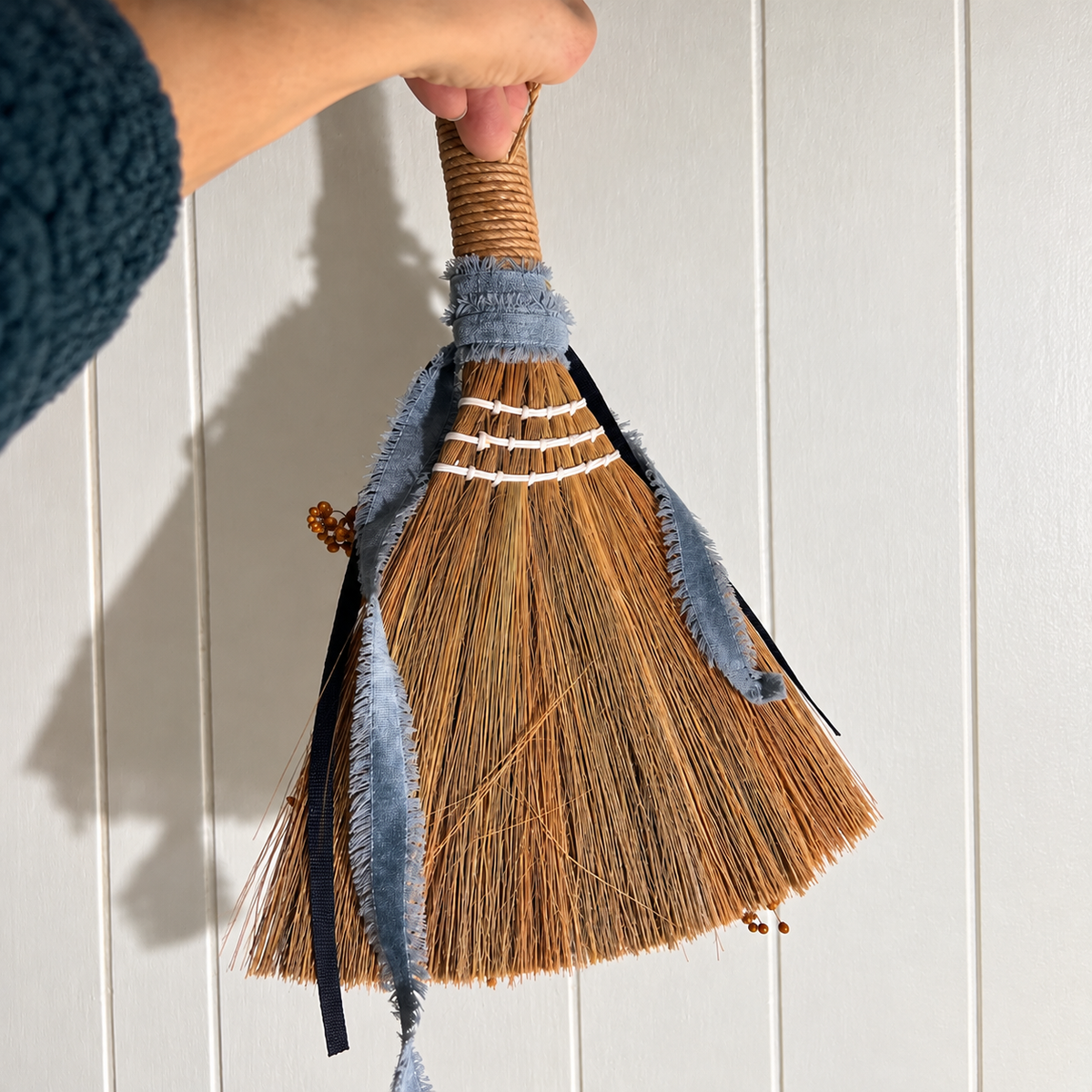 Back view of handmade botanical broom showing natural straw bristles, white stitching, wrapped handle, and trailing blue fabric ribbons.