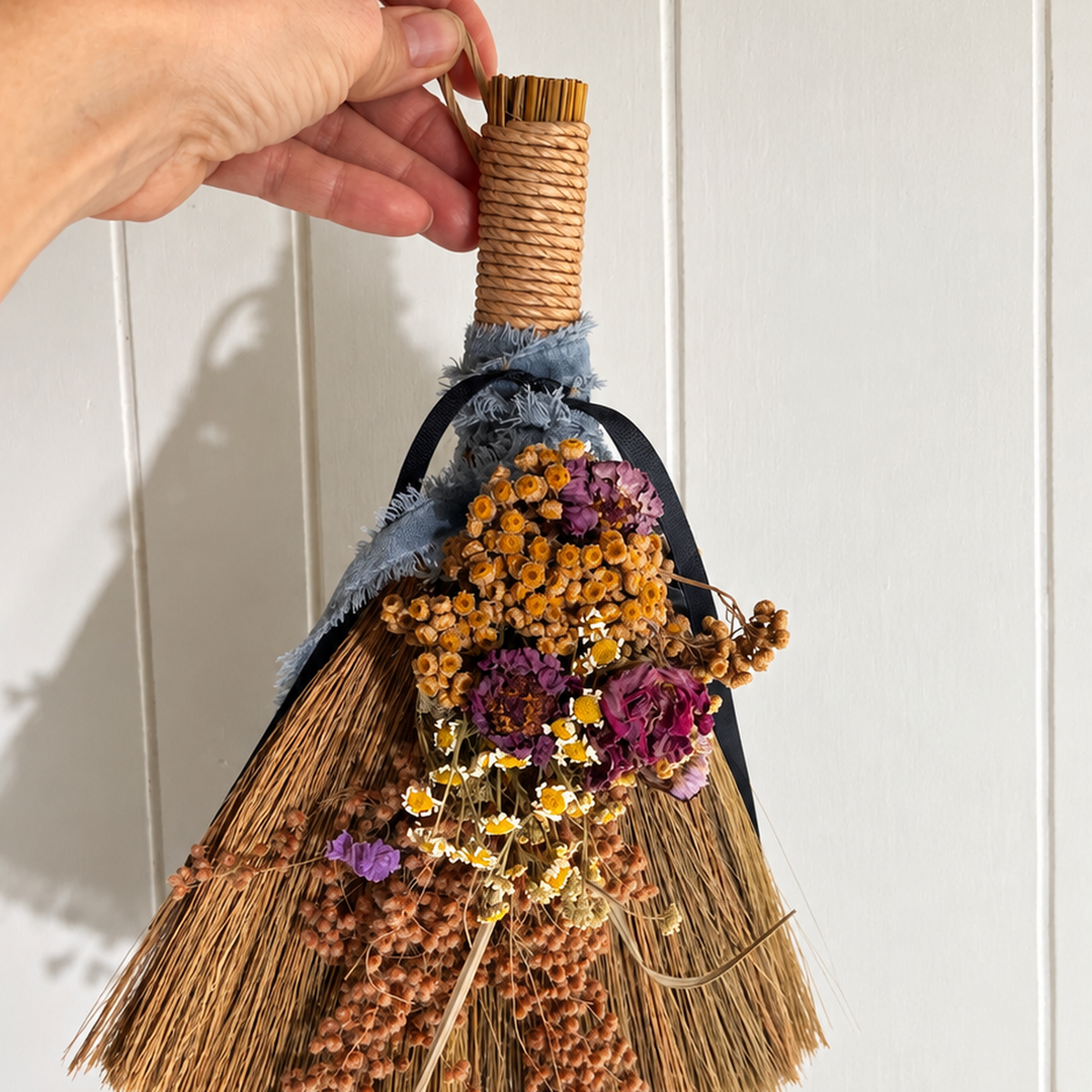 Hand holding a small decorative botanical broom with dried flowers, natural straw bristles, and blue ribbons to demonstrate its size and scale.