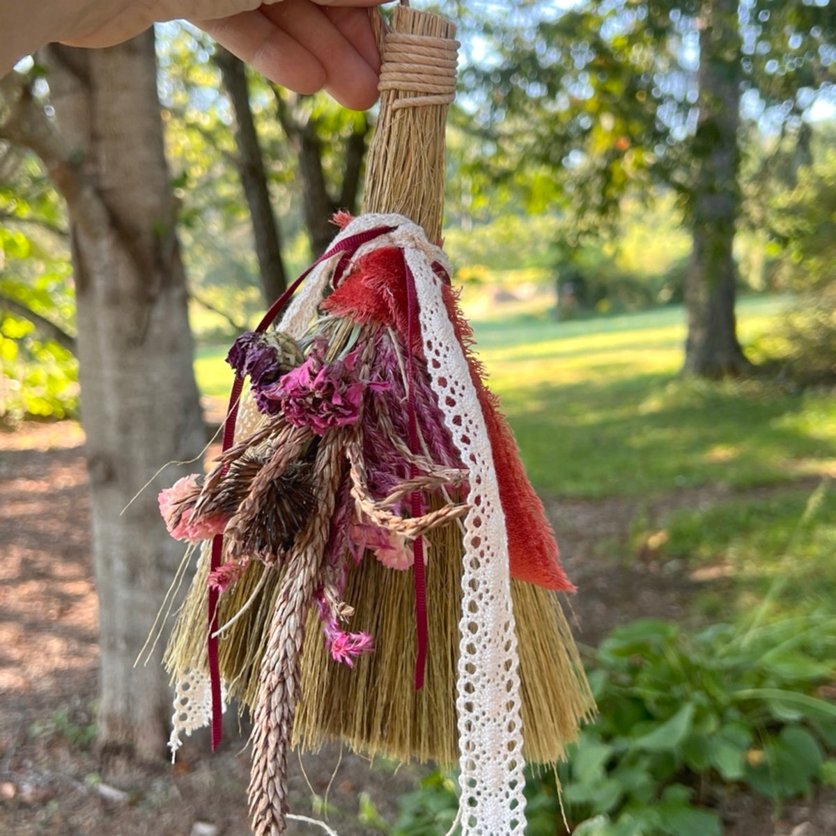 Handmade botanical broom with dried pink flowers, wheat, red ribbon and cream lace held outdoors in a sunlit garden.