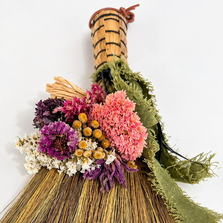 Close-up of a handmade botanical whisk broom featuring pink, purple, yellow and white dried flowers, textured straw bristles and green fabric ribbon.