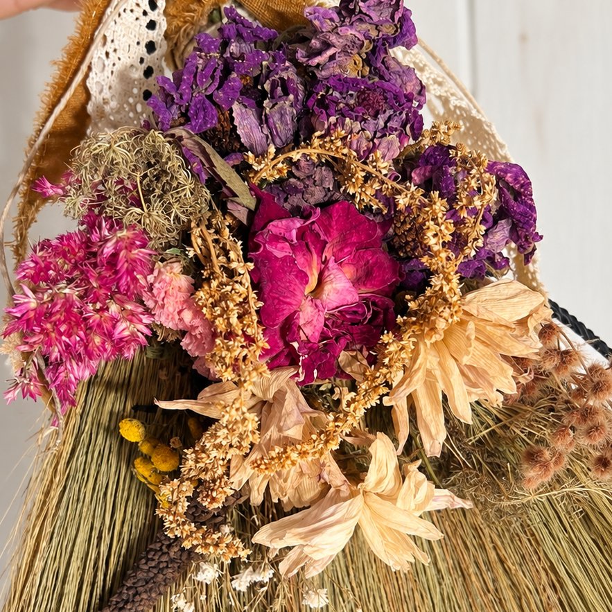 Close-up of dried purple flowers, pink roses, golden botanicals, cream-colored blooms, and vintage lace decorating a handmade straw witch broom.