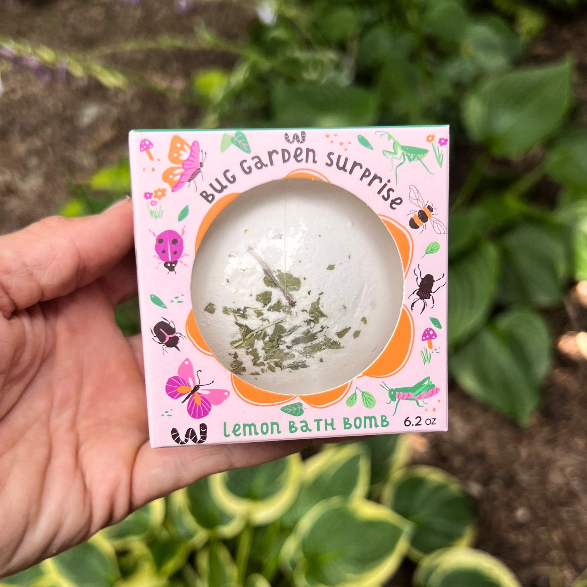 Hand holding the Sow the Magic® Bug Garden Surprise Lemon Bath Bomb gift box outdoors in a lush garden. The windowed package showcases the handmade botanical bath bomb with real peppermint leaves and a hidden bug toy inside, making bath time fun for kids who love nature and insects.