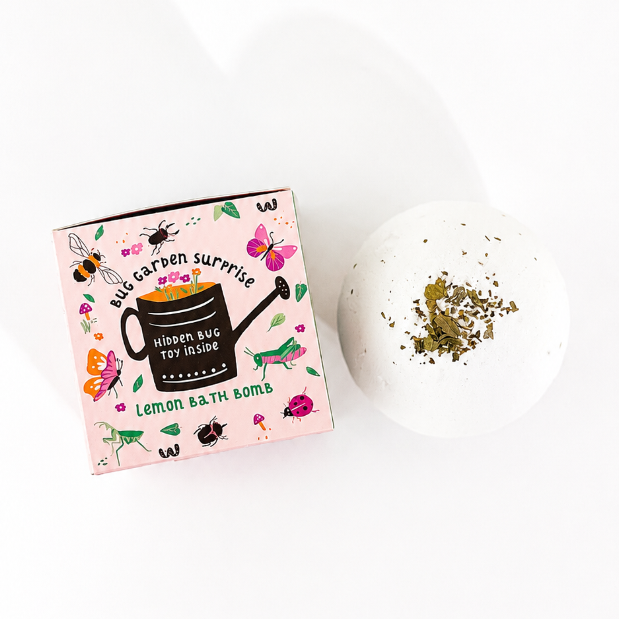 Top-down view of the Bug Garden Surprise Lemon Bath Bomb by Sow the Magic® displayed beside its whimsical bug-themed packaging. The white botanical bath bomb is topped with dried herbs and includes a hidden bug toy surprise, shown on a bright white background.