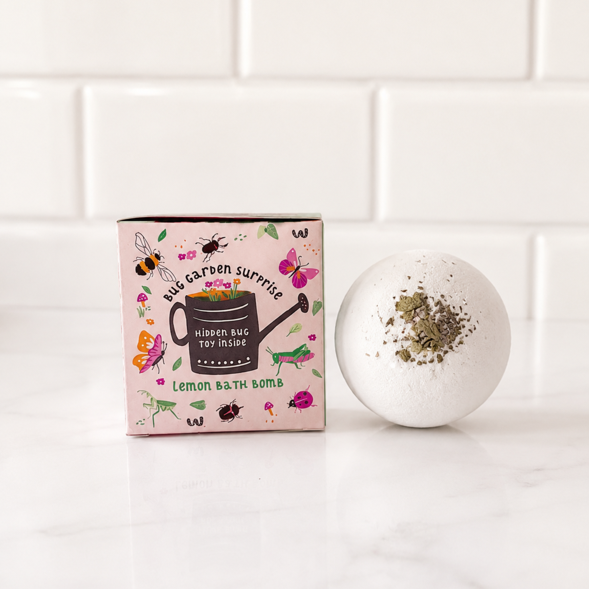 Sow the Magic® Bug Garden Surprise Lemon Bath Bomb displayed beside its colorful gift box. This handmade kids bath bomb is topped with real peppermint leaves and features a hidden bug toy inside. Crafted with shea butter, cocoa butter, and botanical ingredients for fun, fizzy bath time and imaginative play.