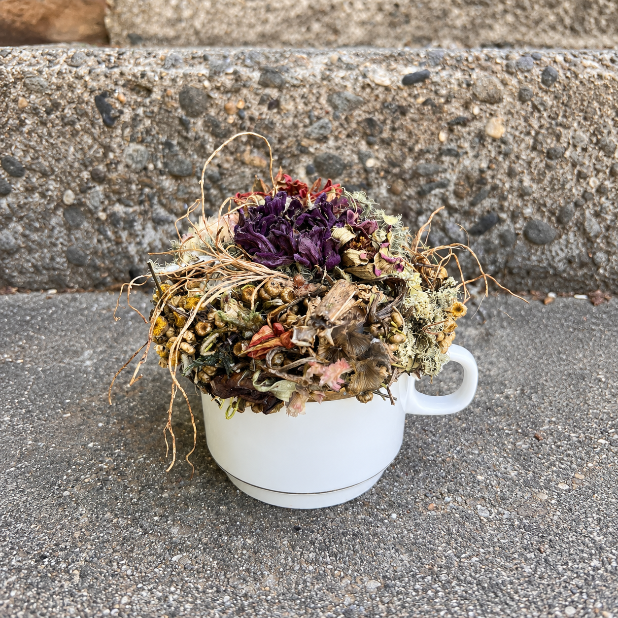 Burgundy and green dried botanical teacup arrangement displayed outdoors on a rustic stone step.