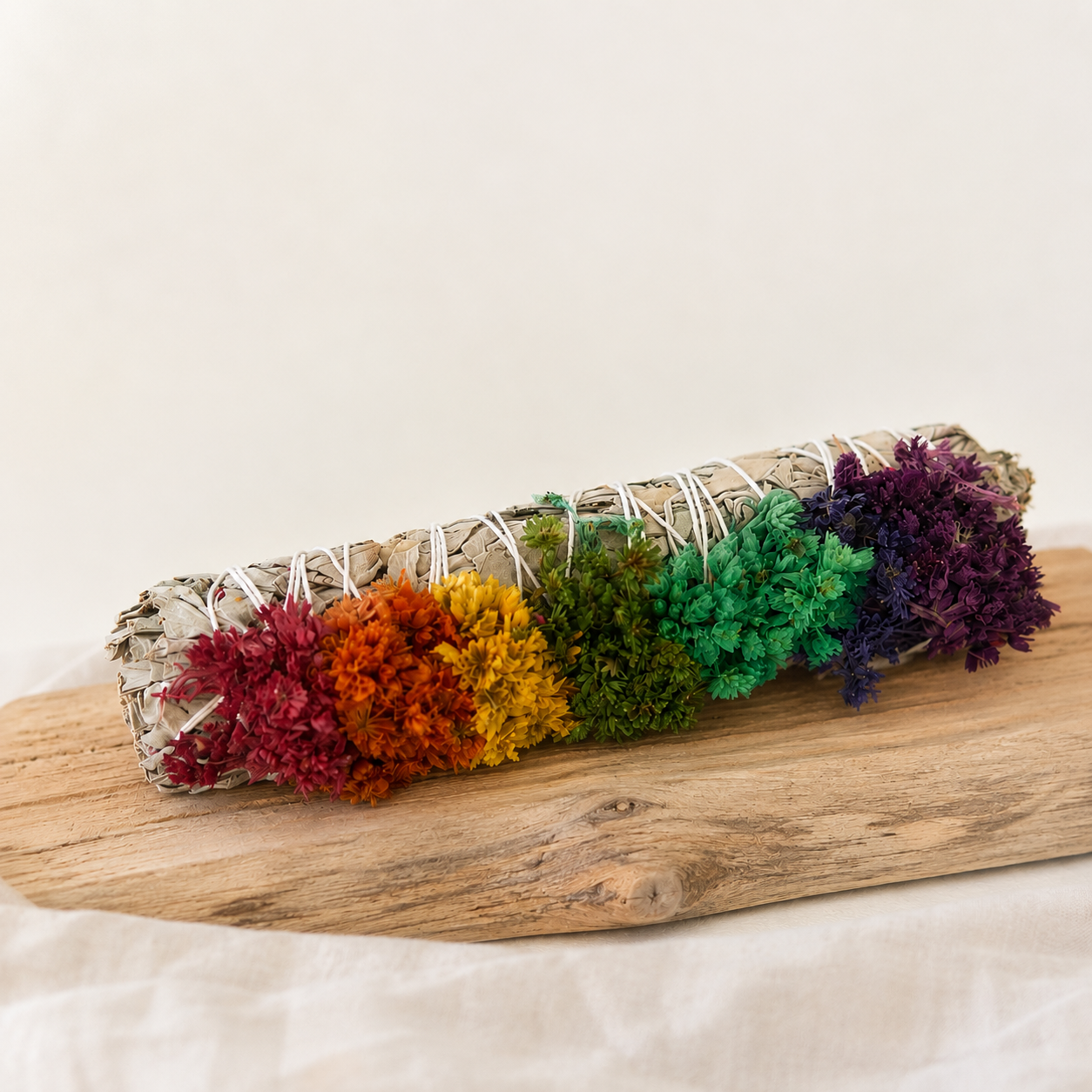 Chakra white sage smudge stick with rainbow sinuata flowers displayed on a natural wood surface