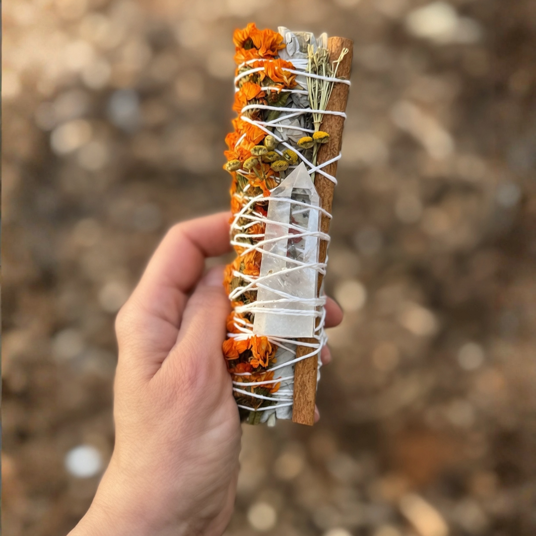 Handcrafted 6-inch White Sage smudge stick featuring a Clear Quartz crystal point, orange sinuata flowers, a cinnamon stick and dried botanicals. Held in hand outdoors with a soft natural background. Handmade by Sow the Magic®