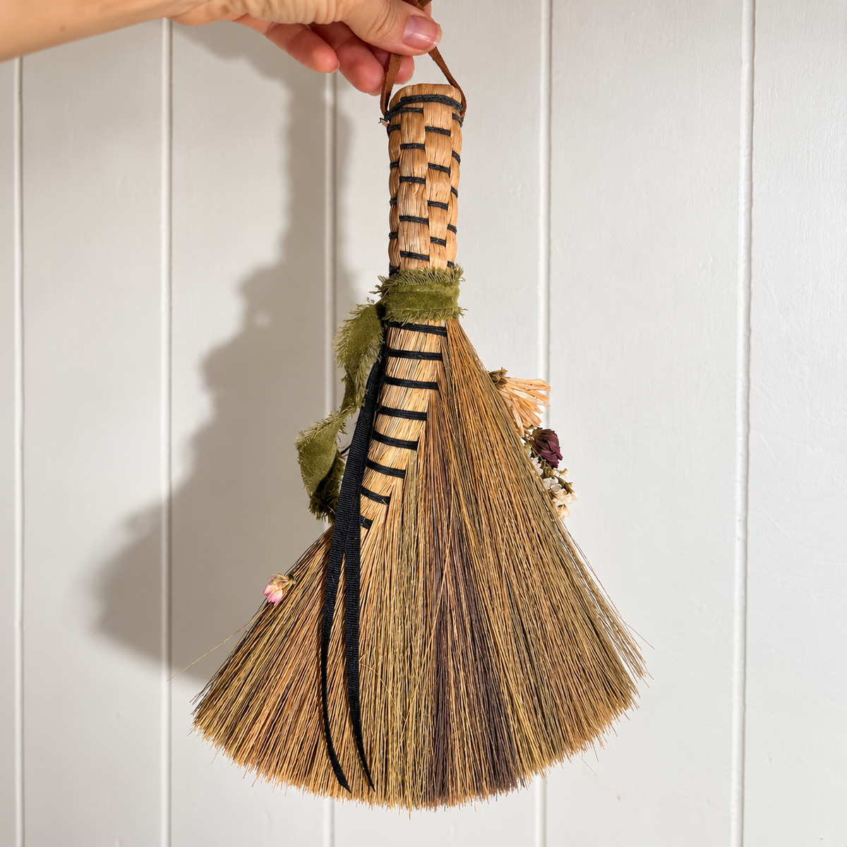 Back view of a handmade botanical broom showing natural straw bristles, black ribbon, woven handle and olive green fabric accents held against white wood paneling.