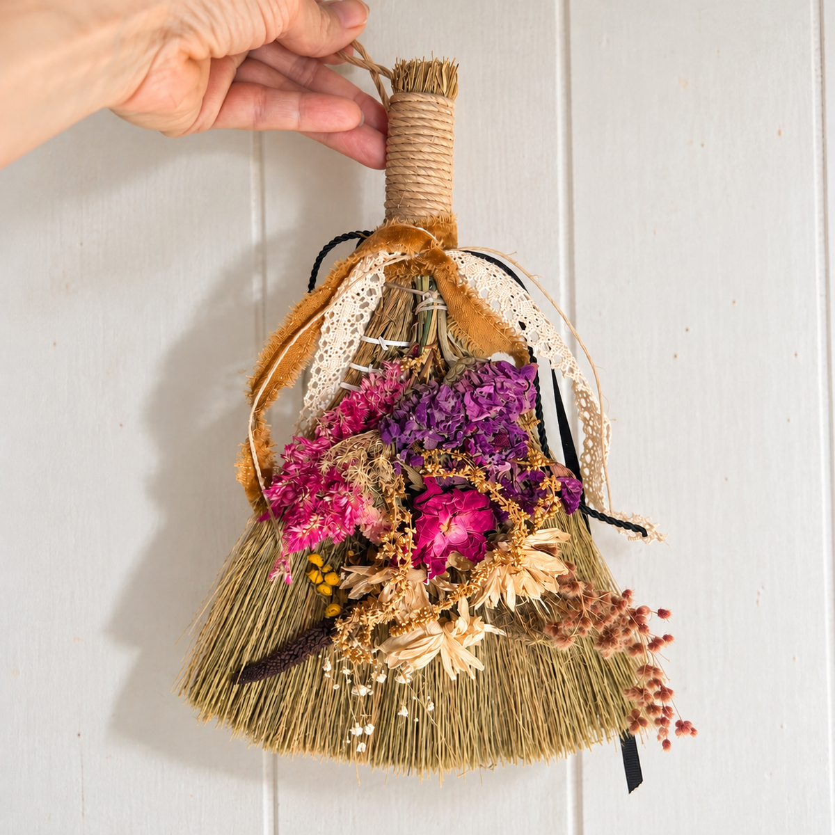 Hand holding a decorative straw broom embellished with dried purple and pink flowers, cream lace, and velvet ribbons against white wood paneling.