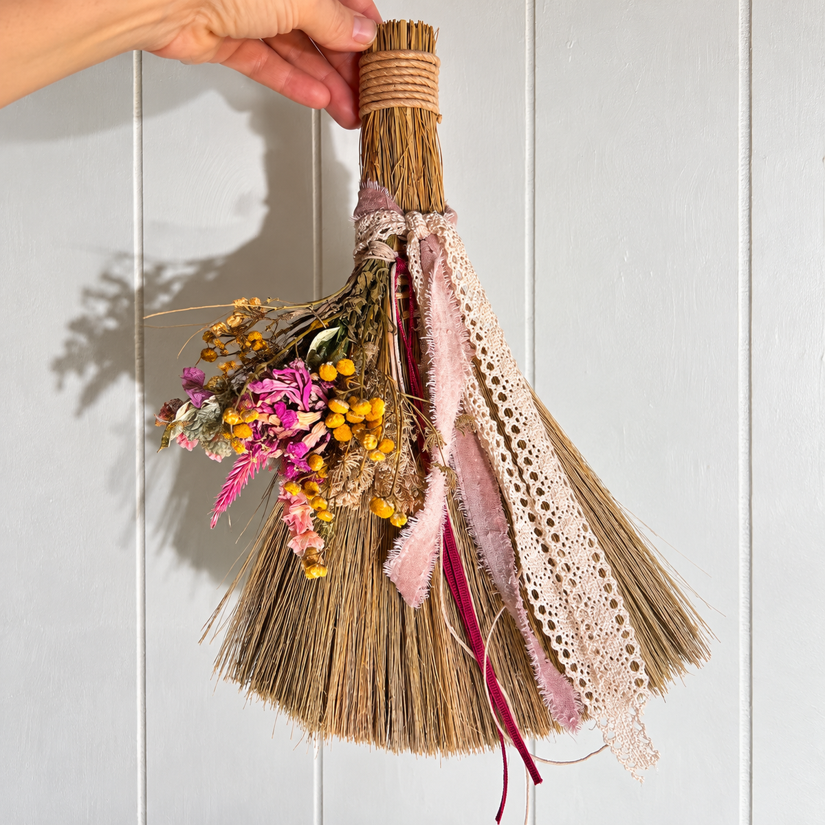 Hand-held decorative botanical broom displaying dried pink and yellow flowers, natural straw bristles, ivory crochet lace and soft blush ribbons against white wood paneling.