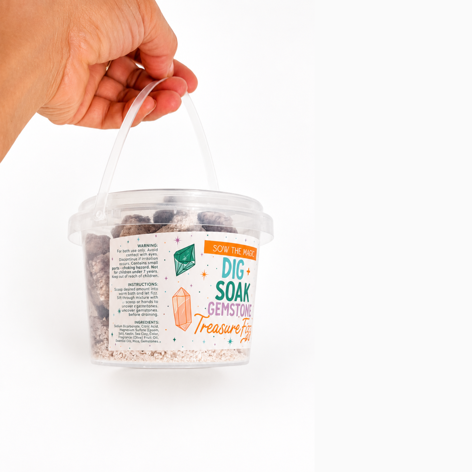 Hand holding the Sow the Magic Dig + Soak Gemstone Treasure Fizz bath bomb bucket by its handle, showing the side label with directions and ingredients. Clear bucket filled with bath bomb nuggets on a bright white background.