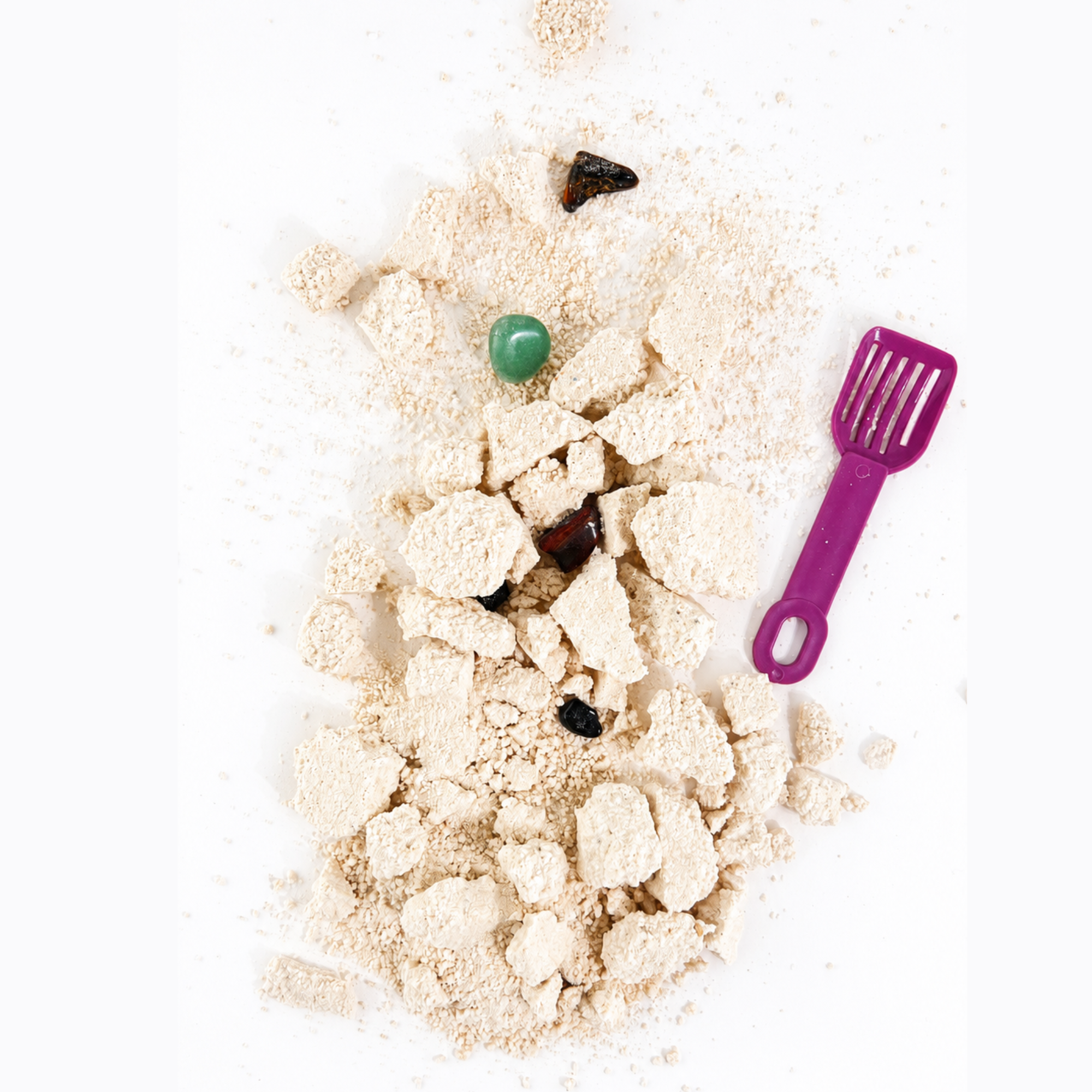 Top-down view of the Sow the Magic Dig + Soak Gemstone Treasure Fizz bath bomb broken into crumbles, revealing colorful polished gemstones alongside a purple digging scoop on a bright white background.