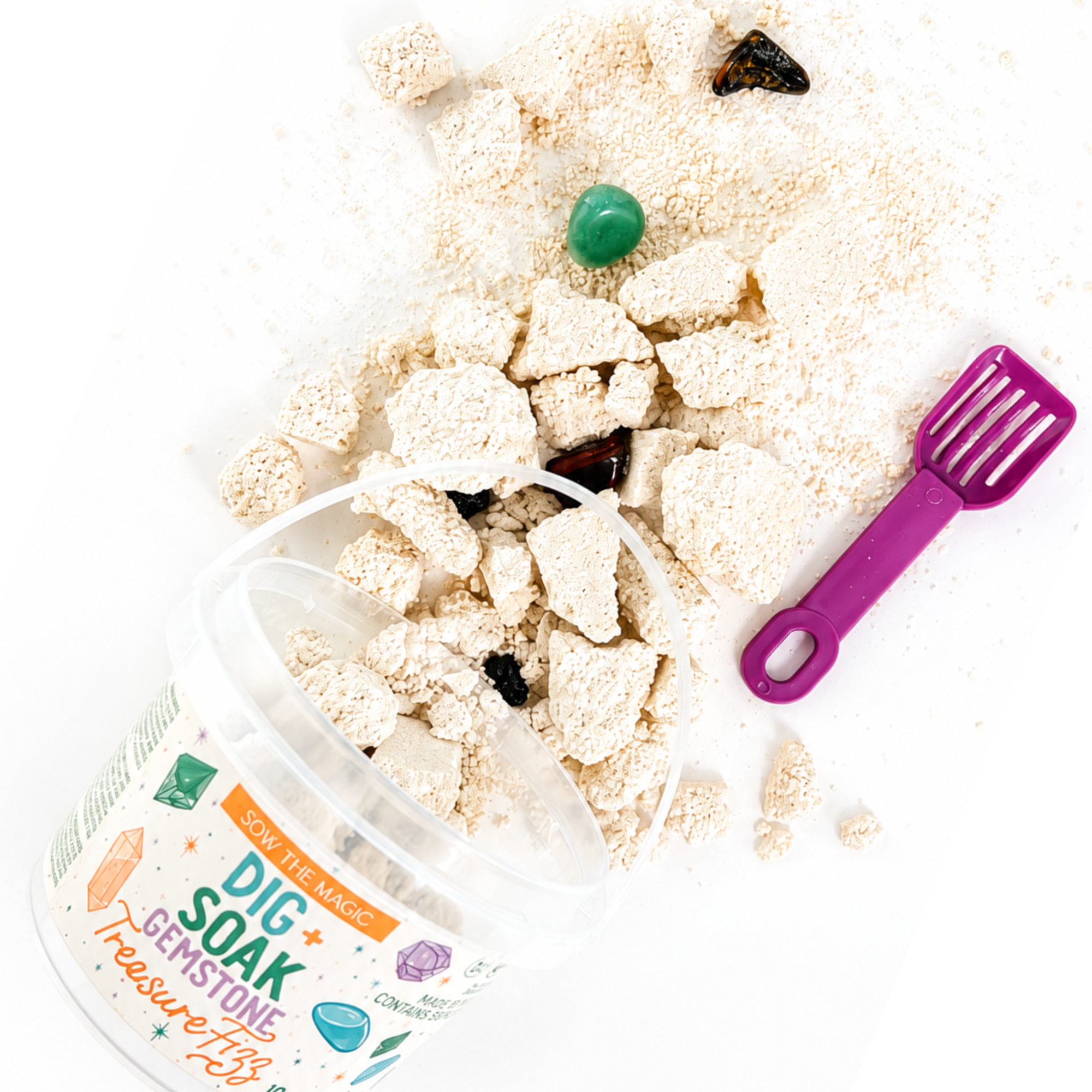 Spilled Sow the Magic Dig + Soak Gemstone Treasure Fizz bath bomb bucket with fizzy bath bomb nuggets, hidden polished gemstones, and a purple digging scoop arranged on a bright white background, showcasing the gemstone discovery experience.