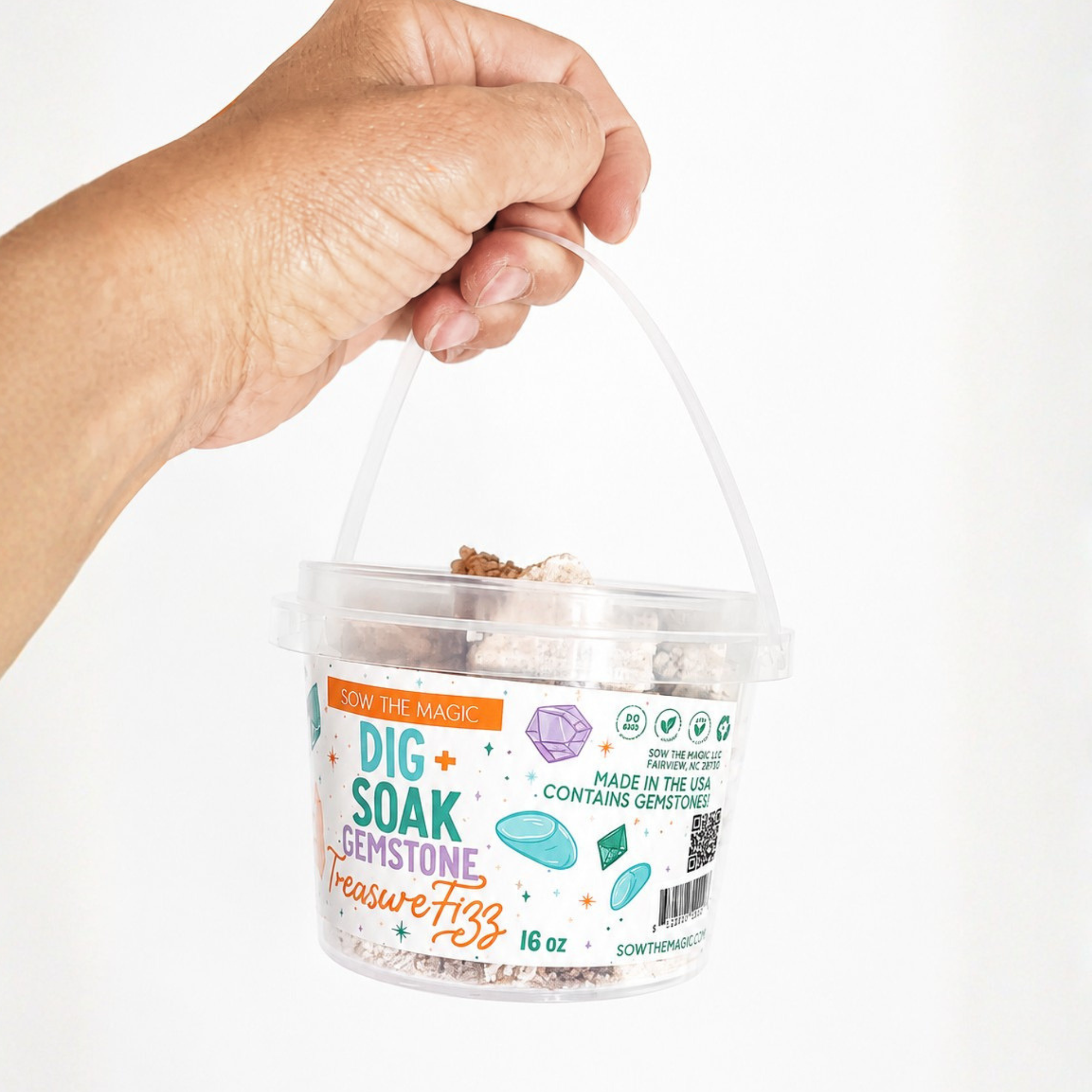 Hand holding the handle of the Sow the Magic Dig + Soak Gemstone Treasure Fizz bath bucket against a bright white background. The clear reusable bucket is filled with fizzy bath chunks that reveal hidden gemstones, with the colorful label clearly displayed.