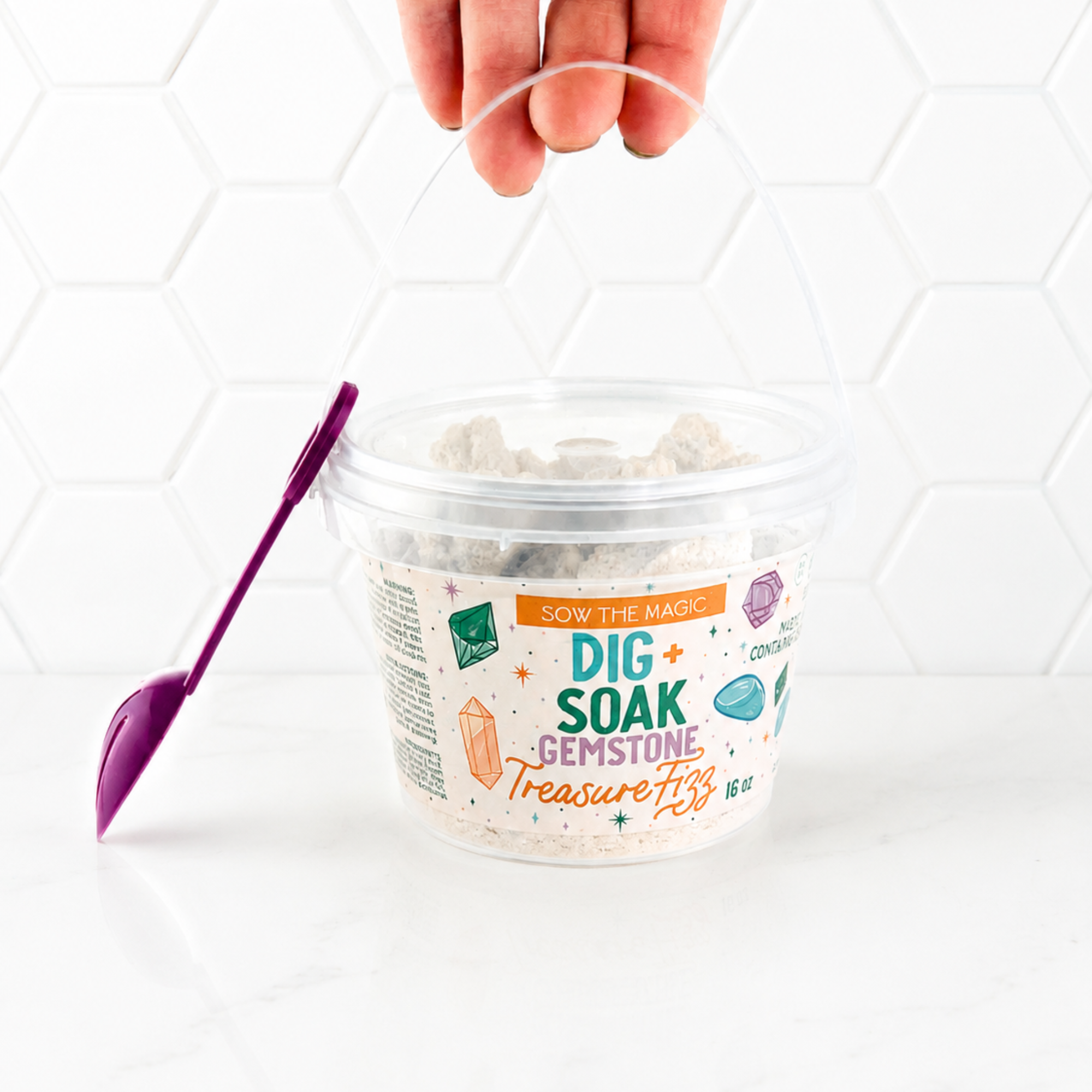 Hand holding the handle of the Dig + Soak Gemstone Treasure Fizz bath bucket by Sow the Magic against a bright white honeycomb tile background. The 16 oz bath fizz bucket includes a reusable scoop and hidden gemstone treasures for a fun sensory bath experience and unique kids' gift.