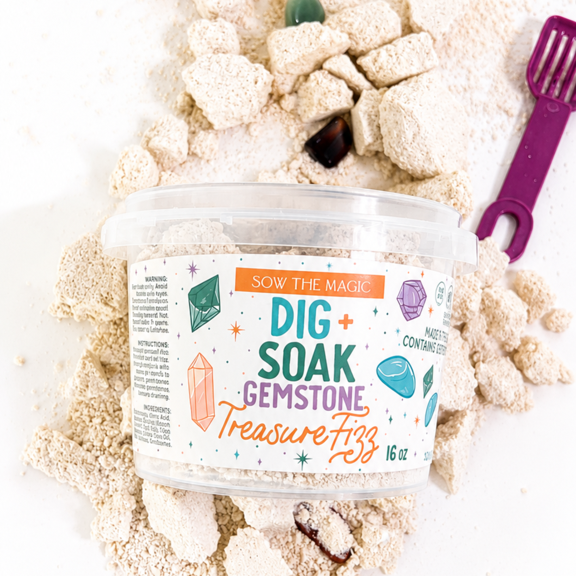 Sow the Magic Dig + Soak Gemstone Treasure Fizz bath dust displayed on a bright white background with scattered fizzy bath chunks, hidden polished gemstones and a purple scoop. The 16 oz bath activity combines a gemstone dig with a colorful fizzing bath experience for kids.