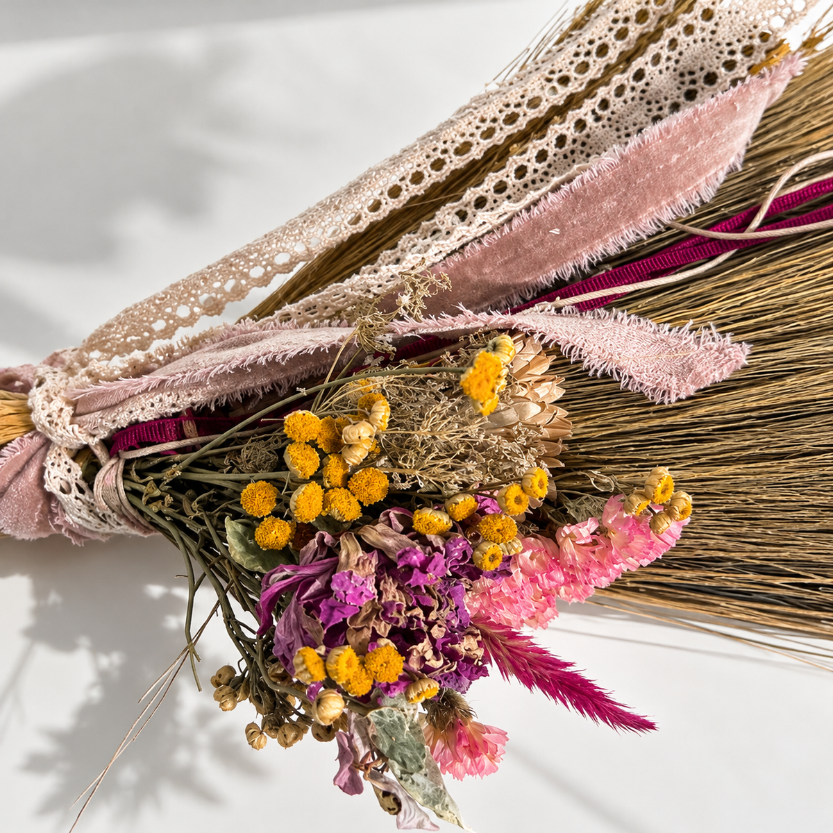 Close-up of handcrafted botanical broom decorations featuring dried yellow button flowers, purple and pink blossoms, delicate ivory lace and frayed blush velvet ribbons.