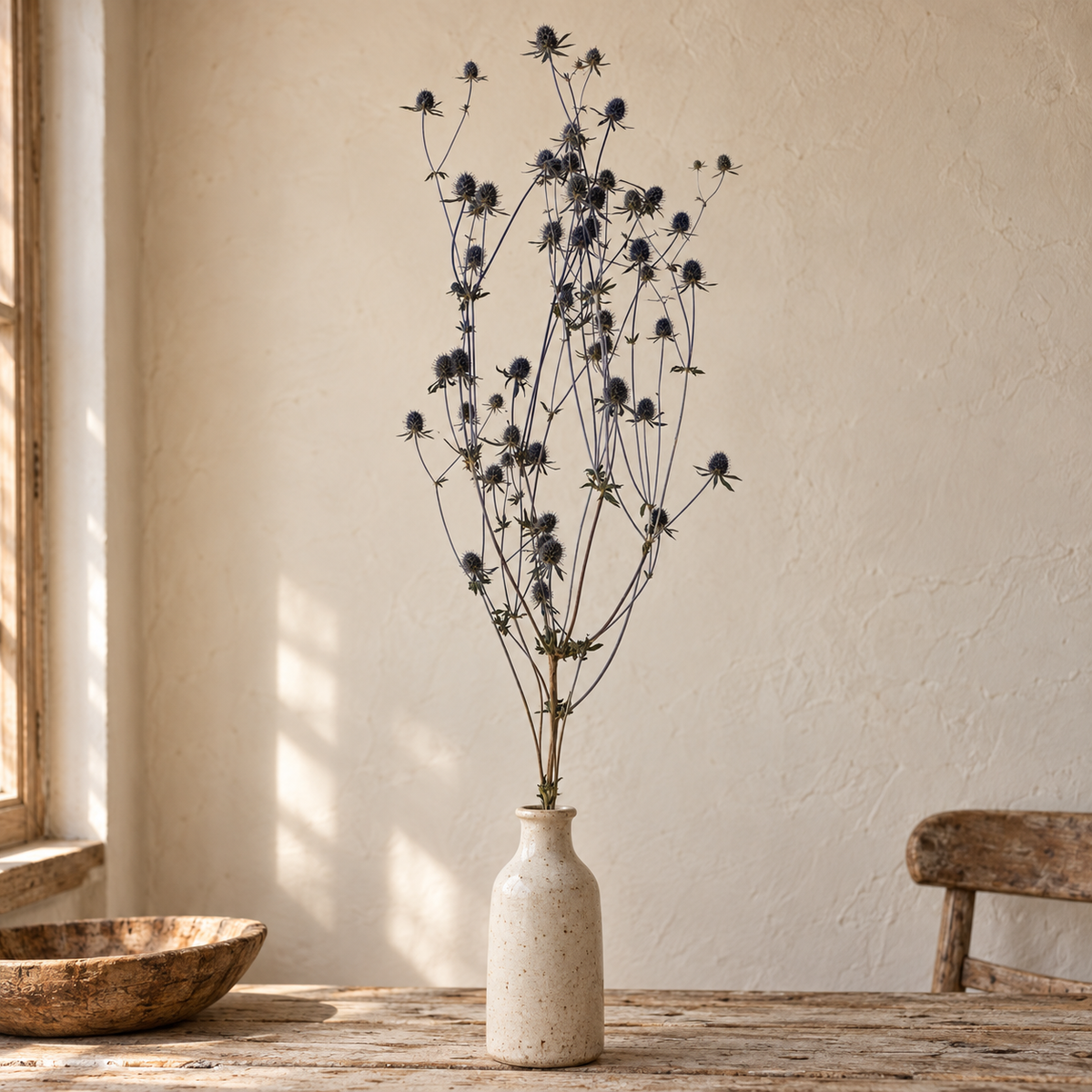 Dried blue sea holly stems with branching flower heads arranged in a cream ceramic vase on a rustic wood table in a warm, sunlit farmhouse setting.