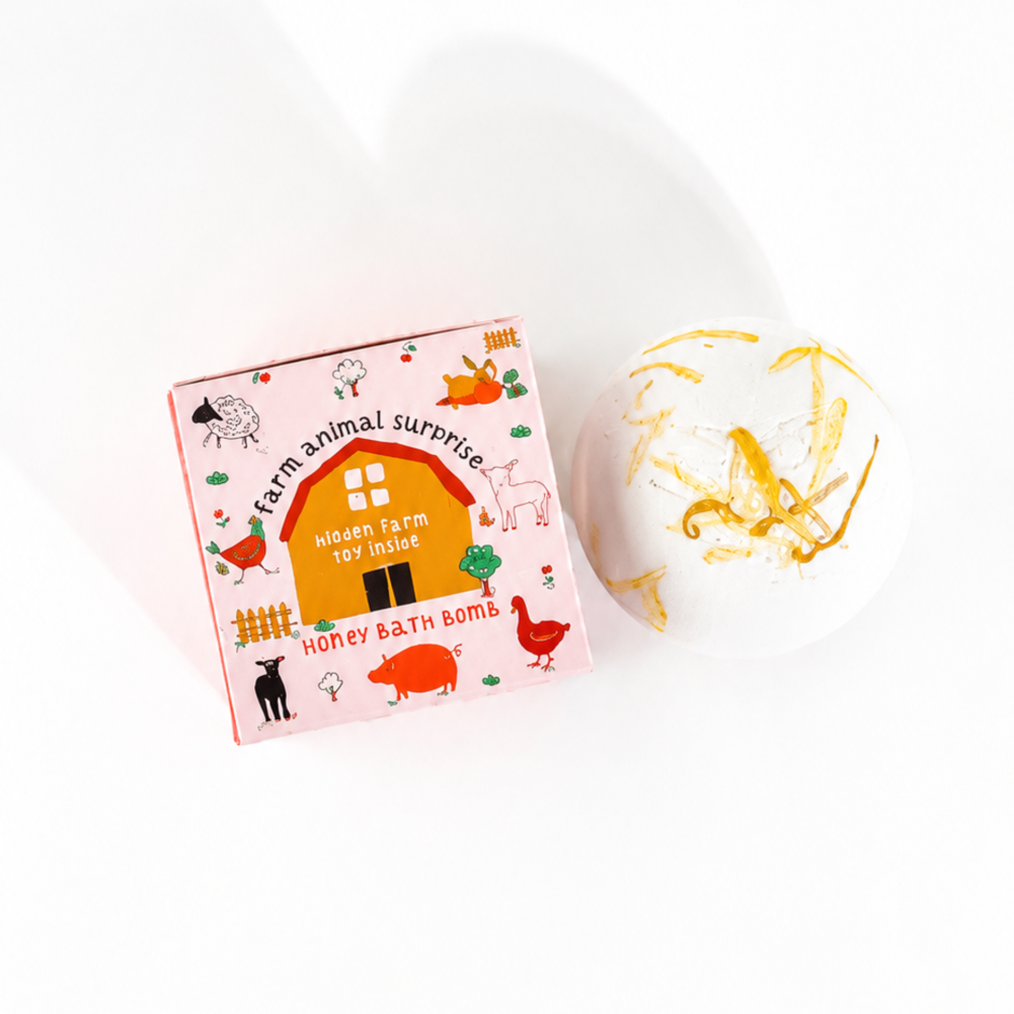 Top-down view of the Farm Animal Surprise Honey Bath Bomb by Sow the Magic® displayed beside its whimsical farm-themed packaging. The white honey-scented botanical bath bomb is decorated with dried calendula petals and includes a hidden farm animal toy surprise, shown on a bright white background.