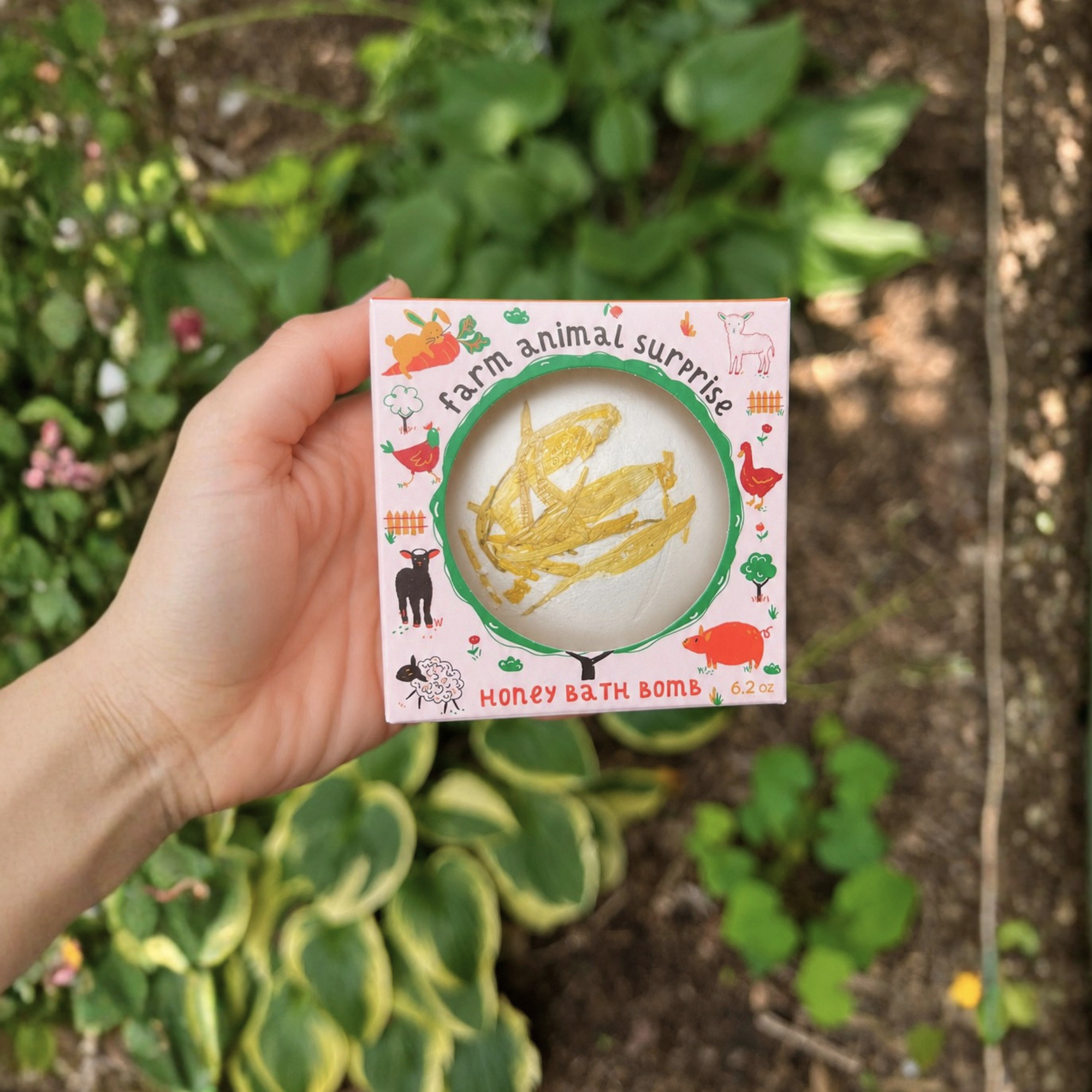 Child-friendly Farm Animal Surprise Honey Bath Bomb by Sow the Magic® in whimsical farm-themed packaging with a window revealing a white honey-scented bath bomb decorated with golden botanicals and a hidden farm animal surprise inside.