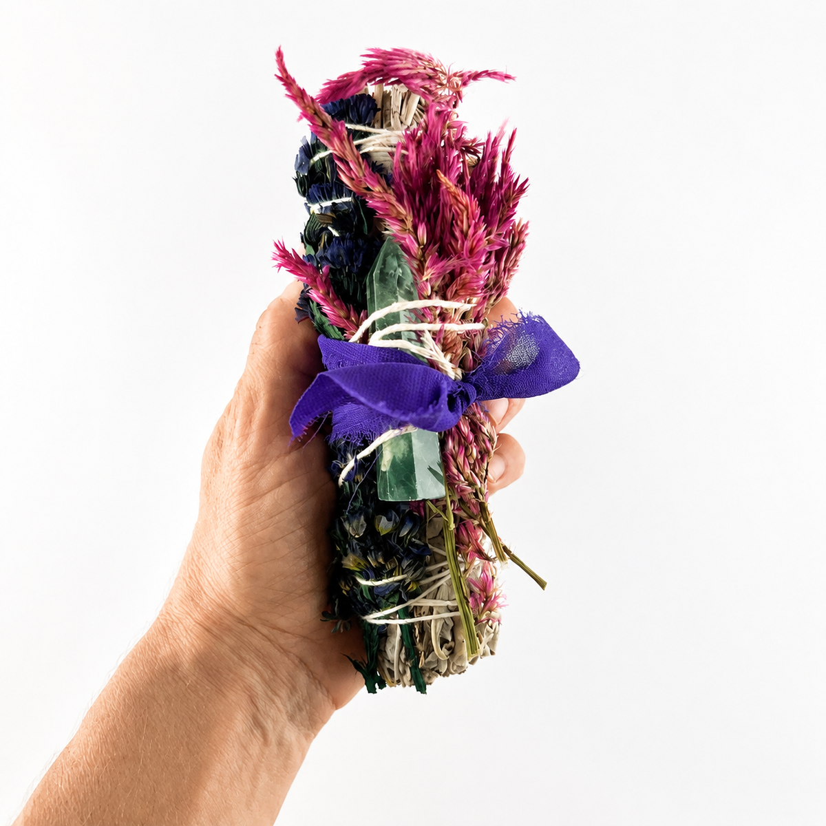 Alt text: Close-up of a hand holding a floral white sage bundle with a fluorite crystal point and purple bow
