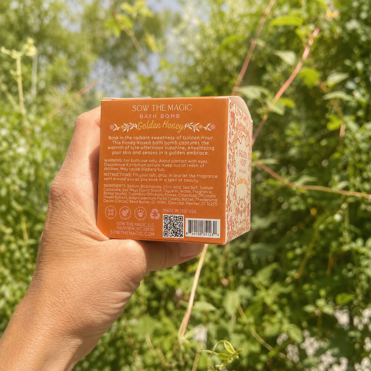 Hand holding the back of the Sow the Magic Golden Honey bath bomb box in a lush garden with softened natural sunlight, highlighting the product details against vibrant green foliage and a warm summer botanical backdrop.