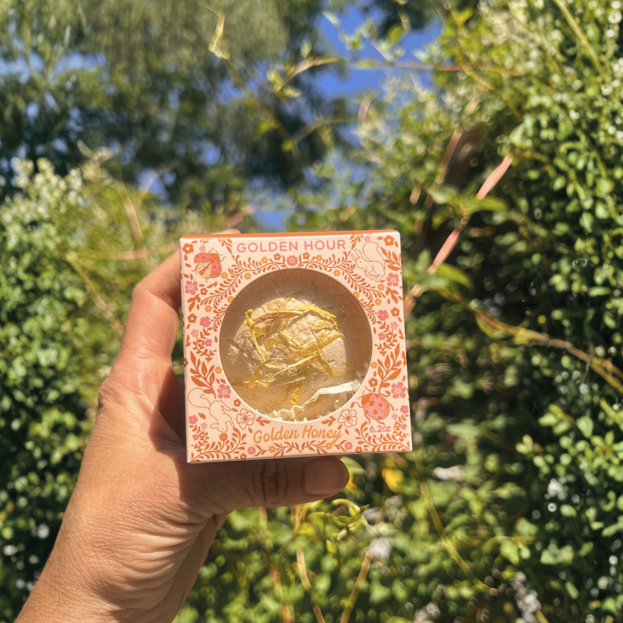 Hand holding the Golden Hour Golden Honey botanical bath bomb gift box in a lush garden with soft natural lighting and blurred greenery in the background.