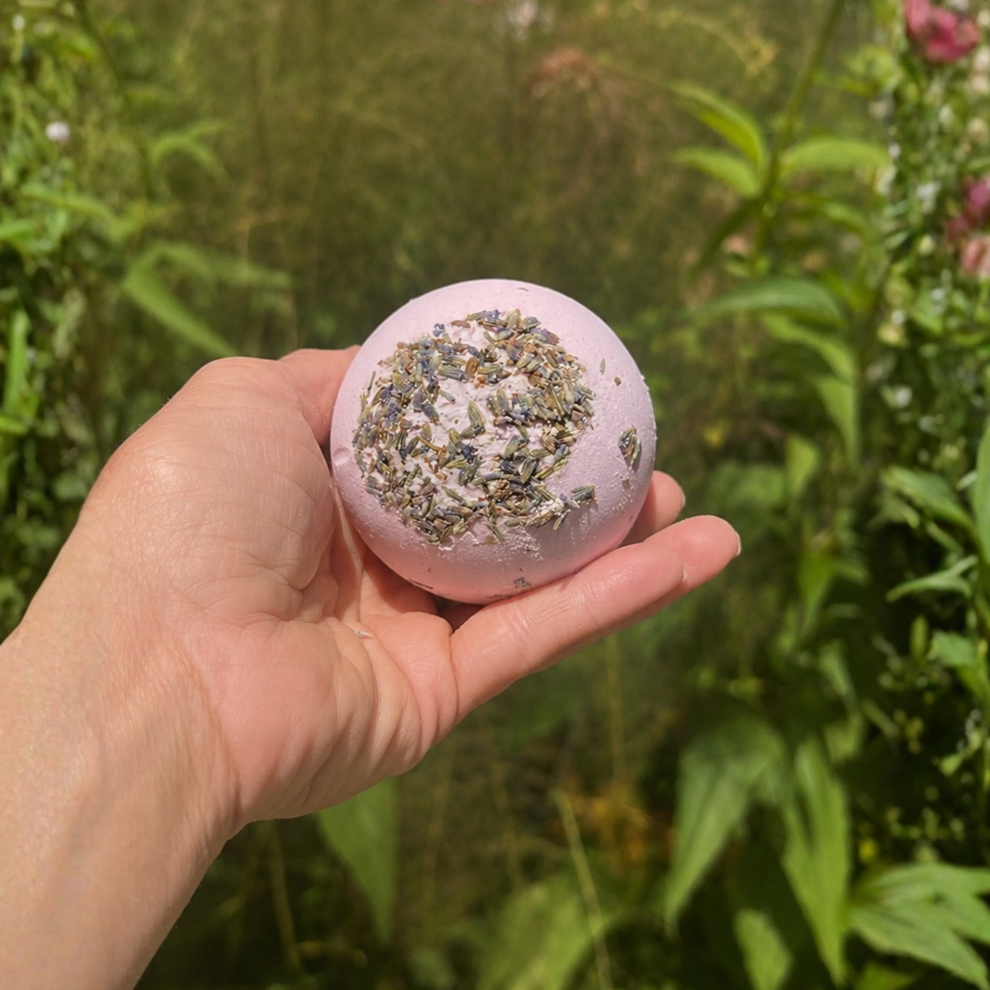Hand holding a Goodnight Moon Lavender Vanilla botanical bath bomb topped with dried lavender buds in a lush garden setting with soft natural lighting and blurred greenery.