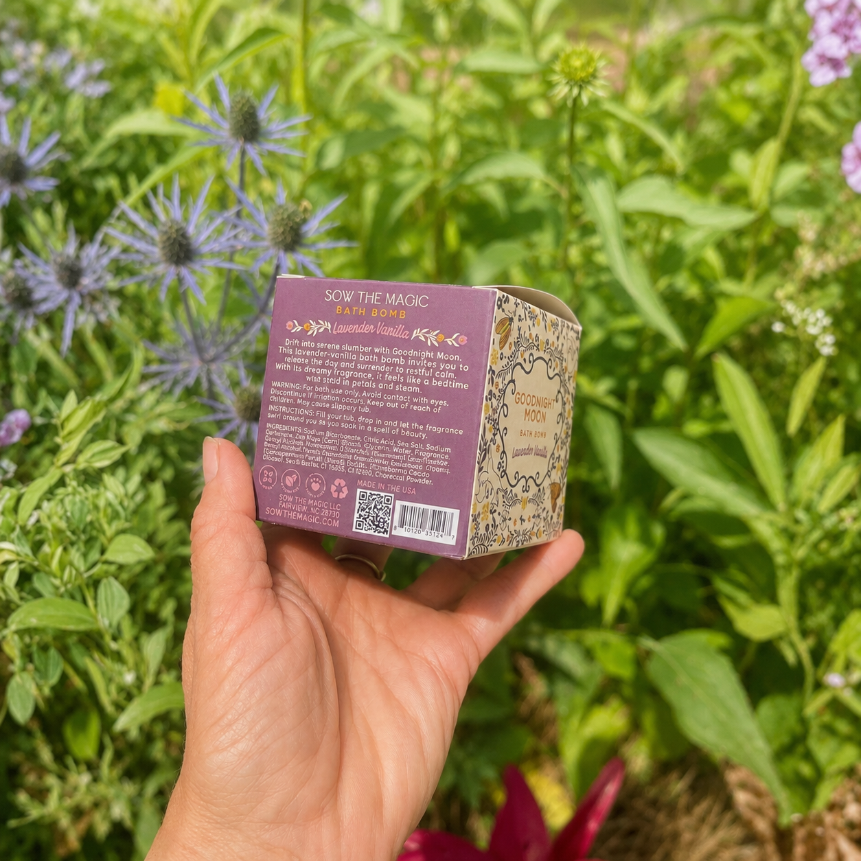 Hand holding the back of the Sow the Magic Goodnight Moon Lavender Vanilla bath bomb box in a lush flower garden with softened natural sunlight, showcasing the product details against blooming blue and purple flowers and vibrant green foliage.