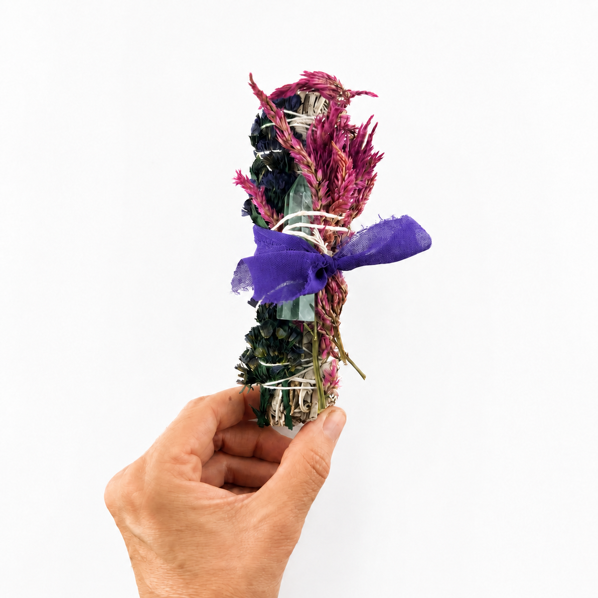 Hand presenting a six-inch white sage bundle decorated with celosia flowers, fluorite and a purple ribbon