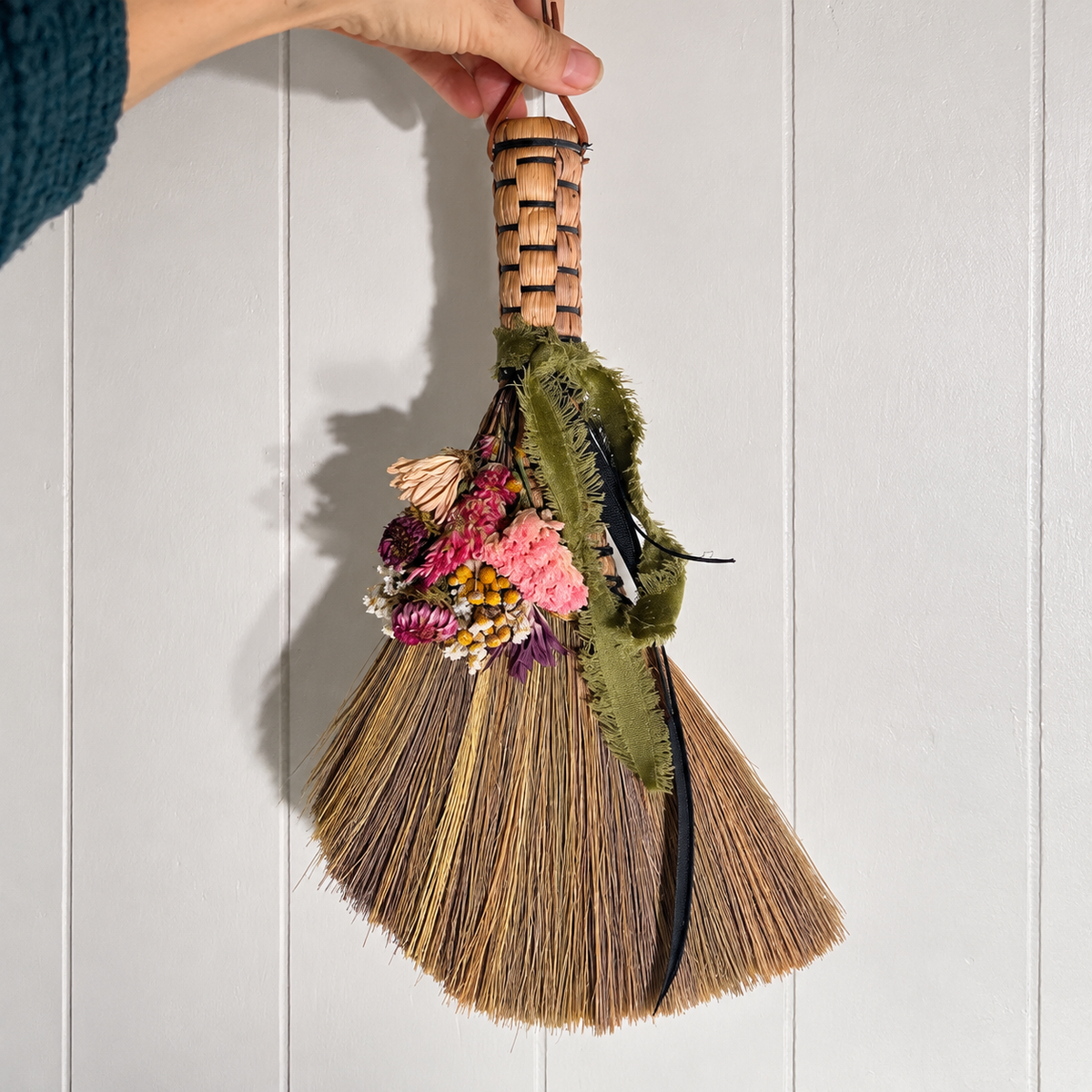 Hand holding a handmade decorative botanical whisk broom with colorful dried flowers, olive green ribbon and natural straw against white wood paneling.