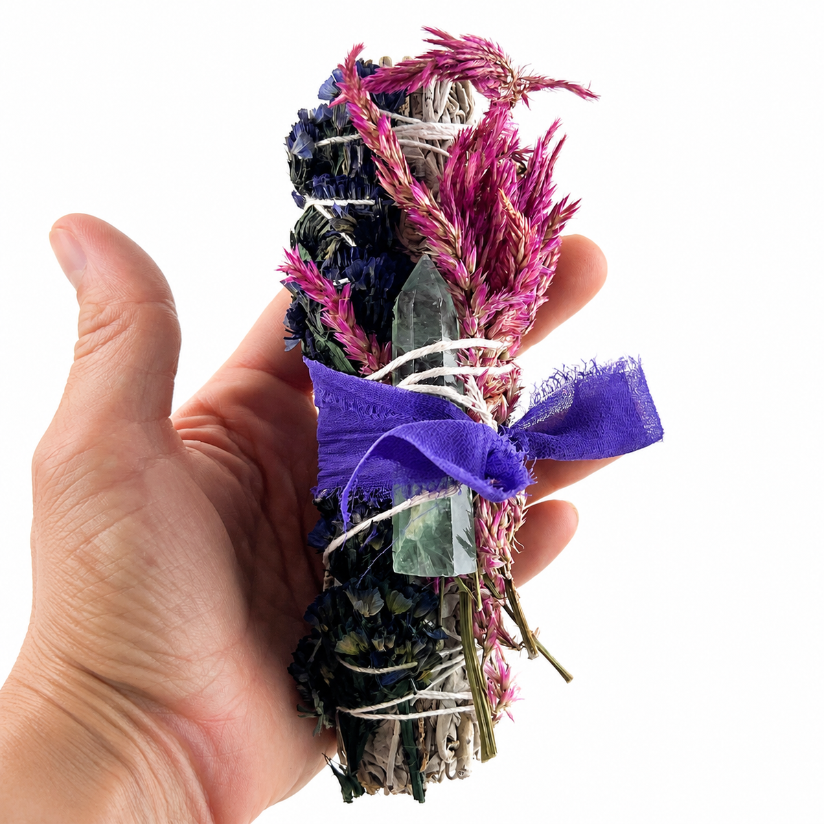 Hand holding a white sage bundle adorned with celosia flowers, blue botanicals, fluorite and purple ribbon