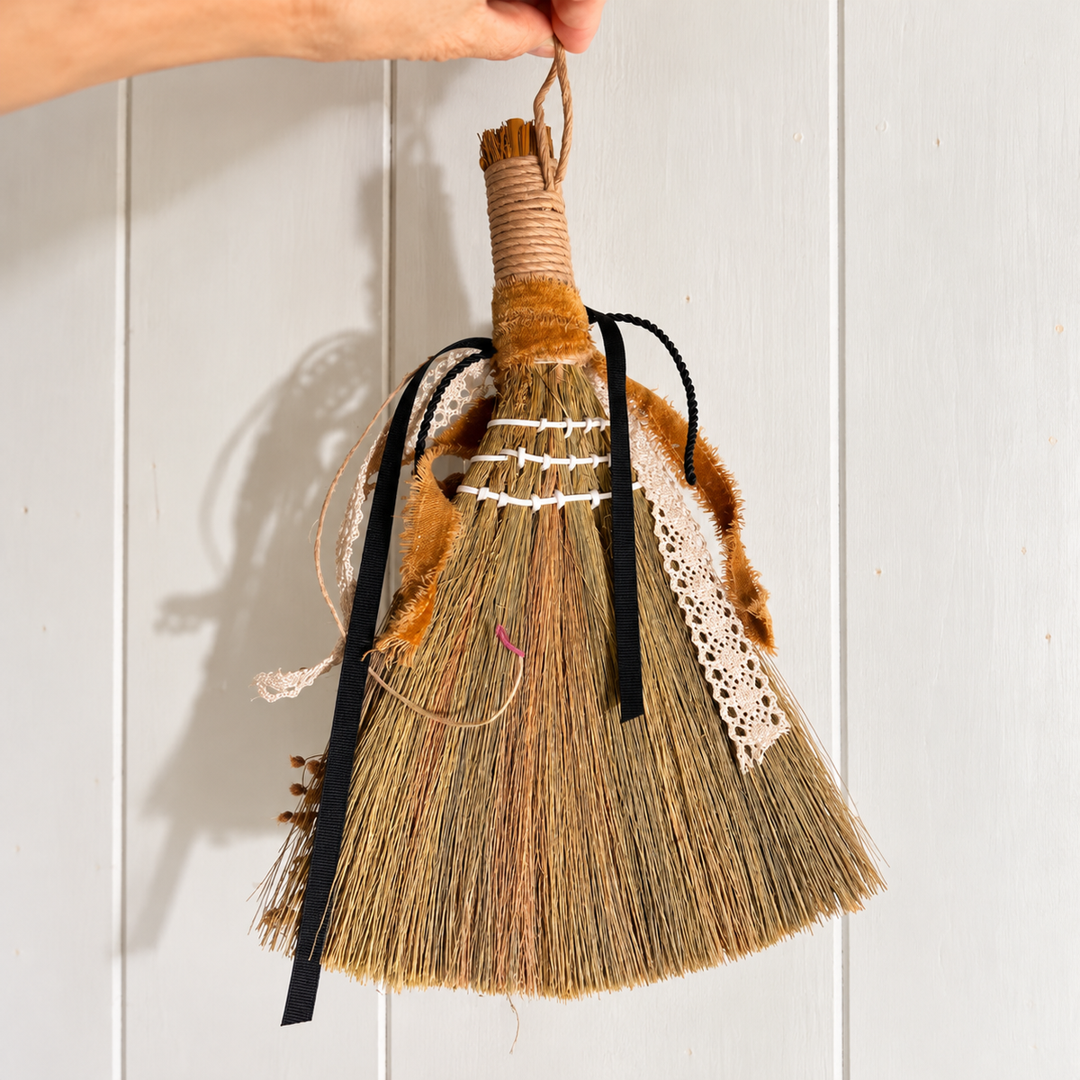 Back view of a handmade botanical broom showing natural straw bristles, white stitching, twine-wrapped handle, black ribbons, and vintage lace trim.