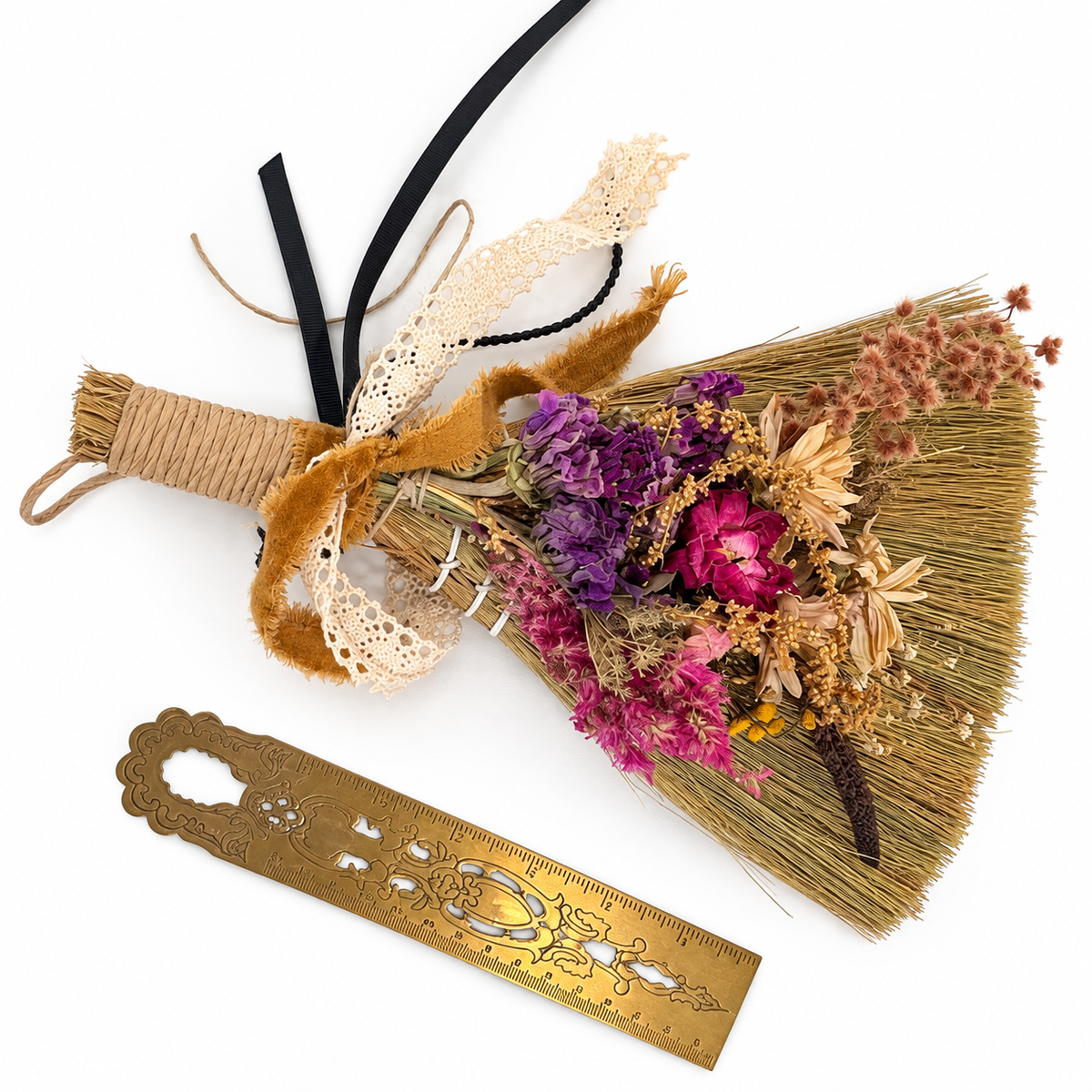 Handmade decorative botanical broom adorned with purple, pink, and cream dried flowers, vintage lace, and ribbons, displayed beside a brass ruler for size reference.