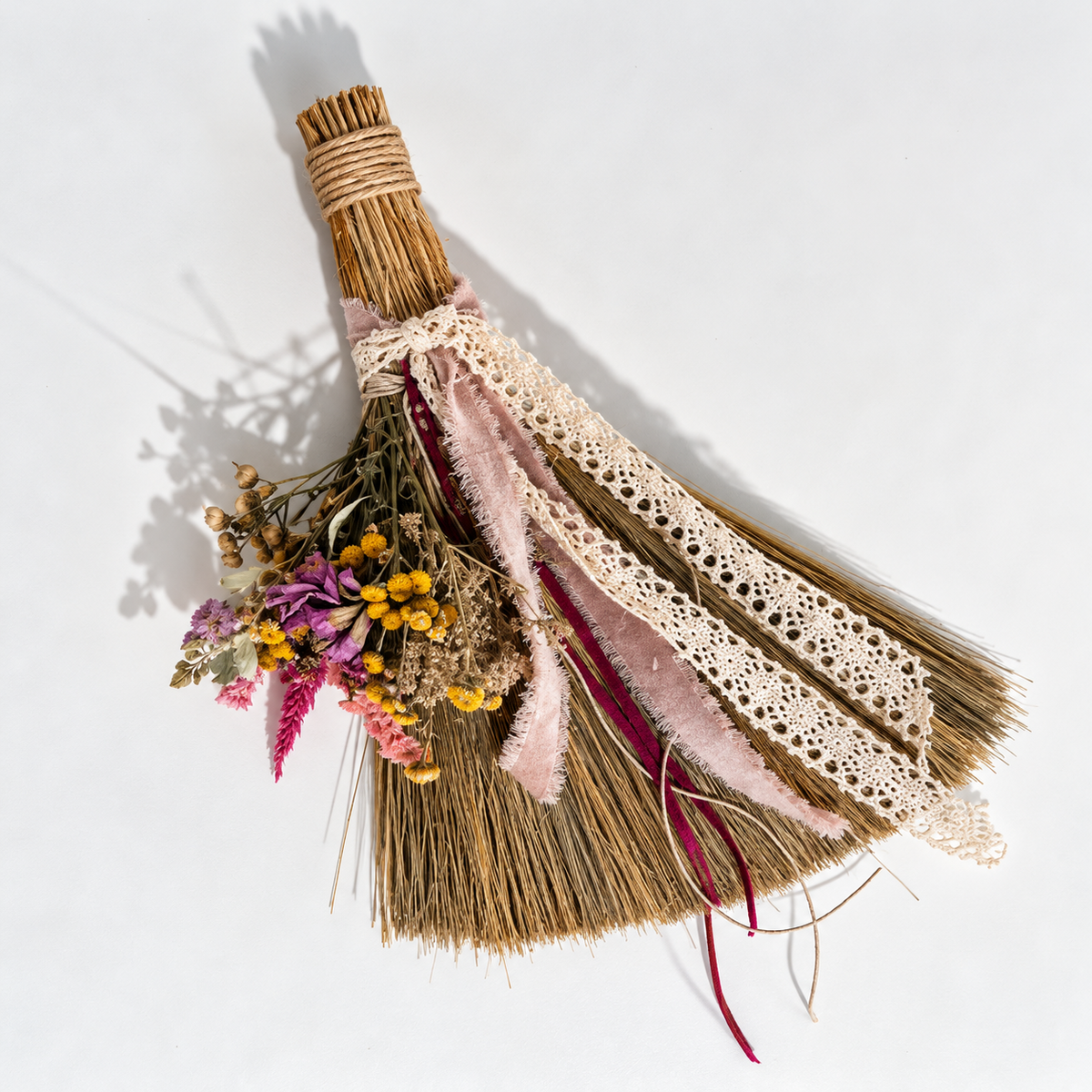 Handmade decorative botanical broom featuring dried pink, purple and yellow flowers, vintage ivory lace, blush velvet ribbons and natural straw on a white background.