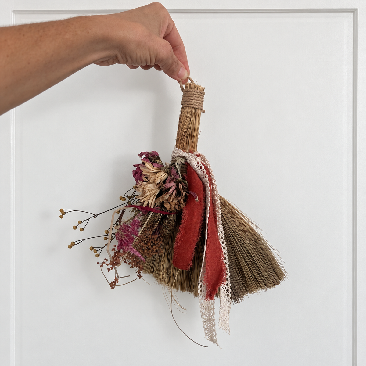 Hand holding The Hearthkeeper’s Botanical Broom to show its hanging size, dried flowers, red ribbon and cream lace.