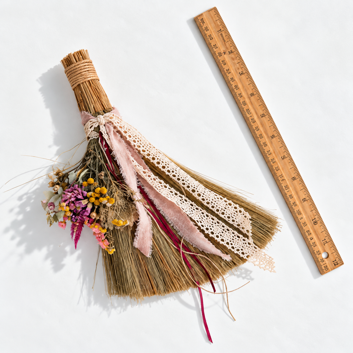 Handmade mini botanical broom with dried flowers, ivory lace and blush ribbons photographed beside a 12-inch wooden ruler to show its size and proportions.