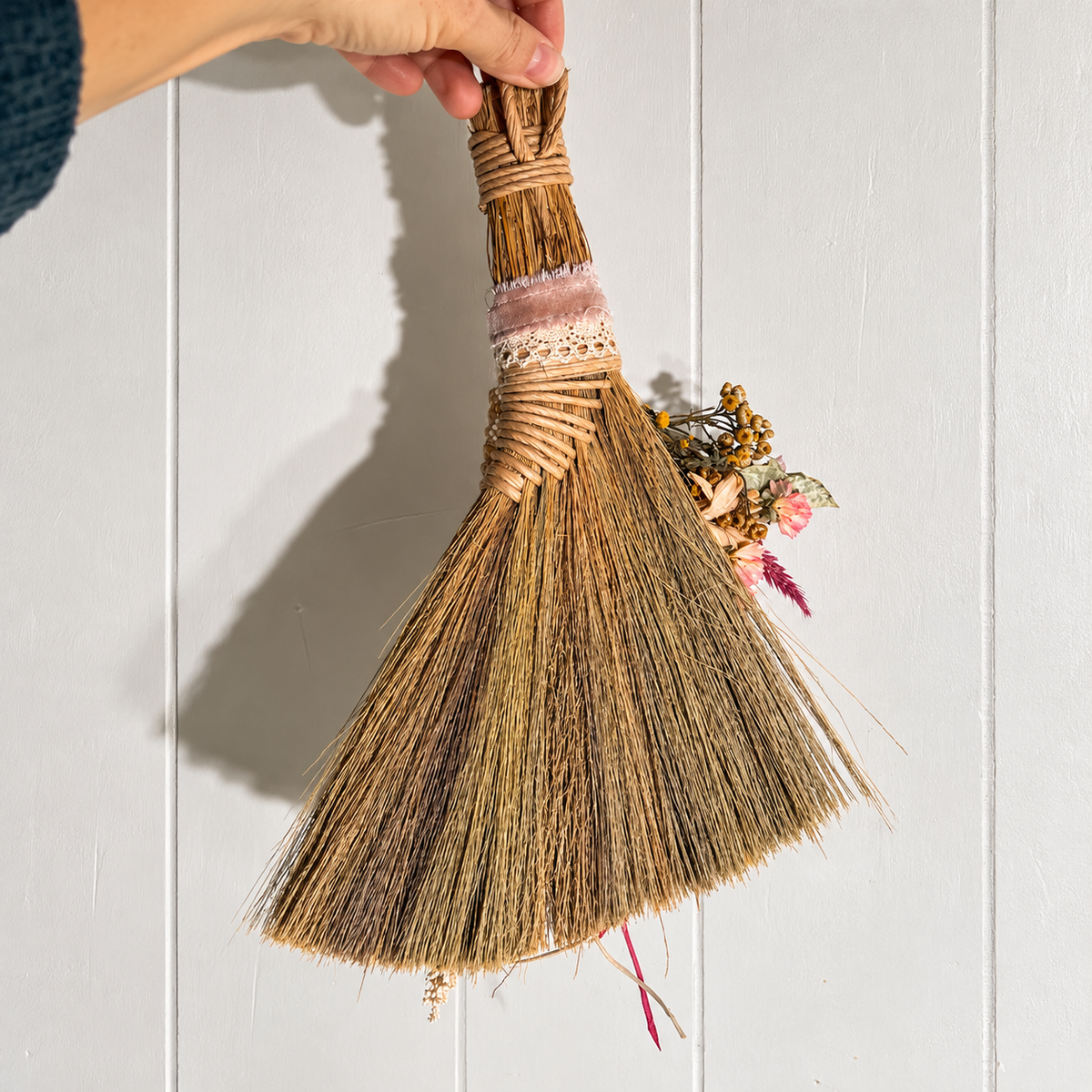 Back view of a handcrafted decorative straw broom with natural twine-wrapped handle, dried botanical accents and trailing ribbons, held against white wood paneling.