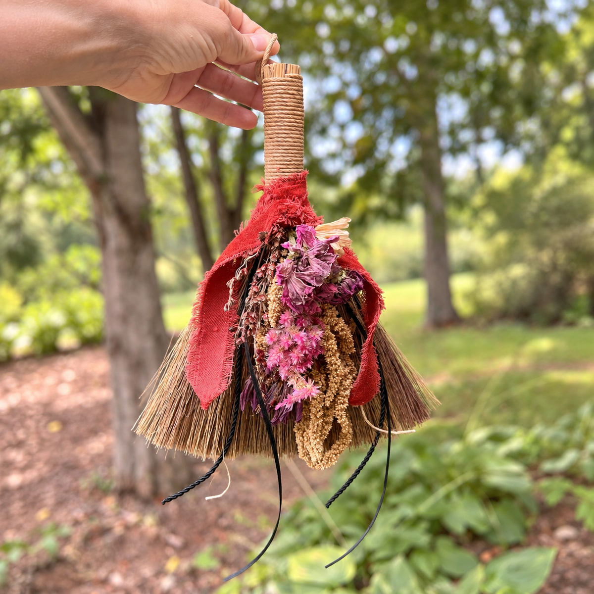 Hand holding the Wildheart Botanical Broom outdoors featuring dried botanicals, rustic ribbon and natural broomcorn